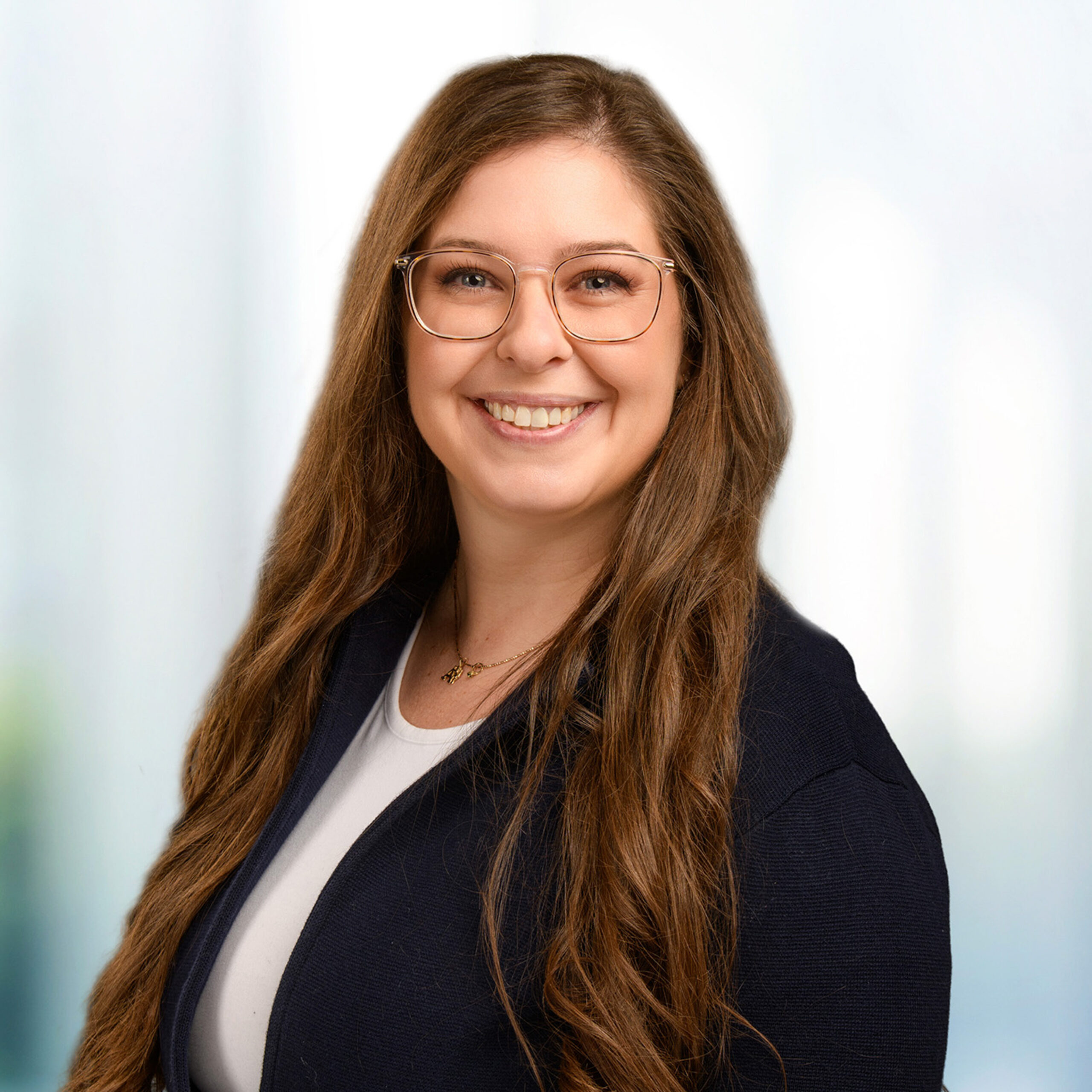 Smiling woman with long brown hair and clear-framed glasses wearing a navy blazer over a white top against a soft light background