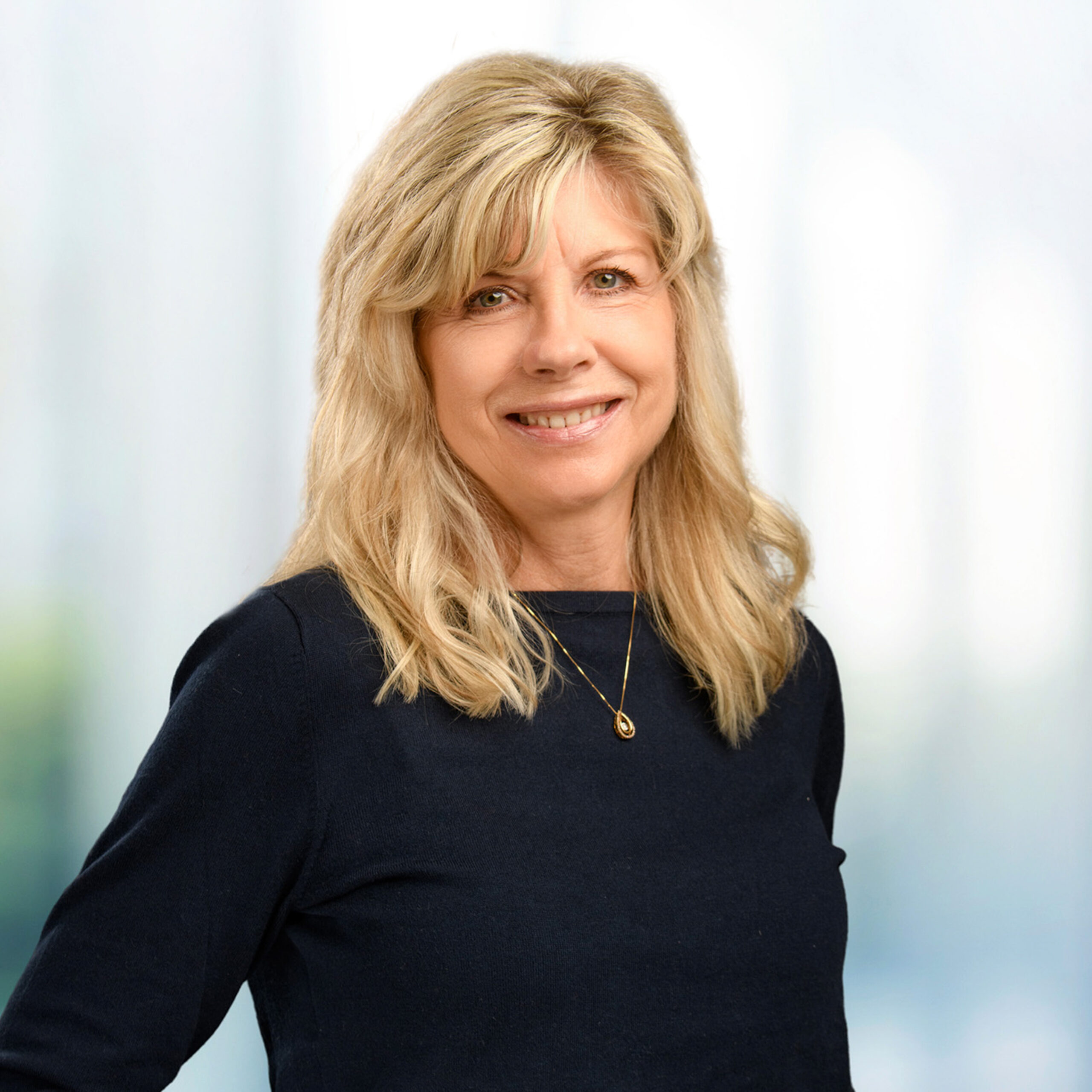 Smiling woman with shoulder-length blonde hair in a navy top and gold pendant, soft blurred background