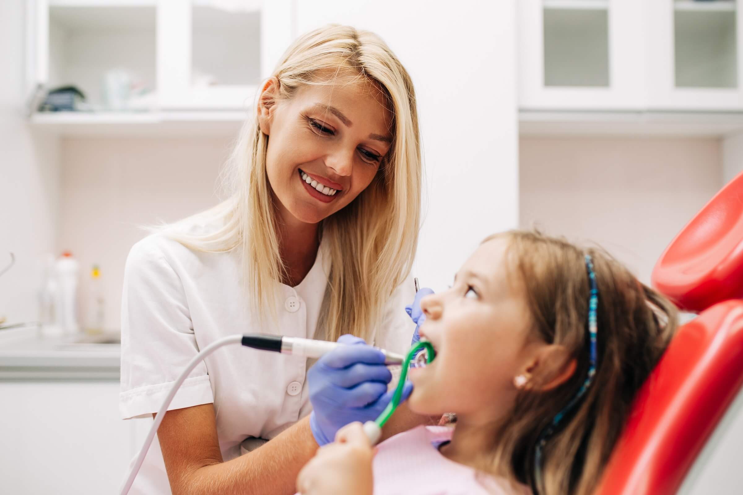 Smiling female dental hygienist in a white uniform and gloves cleaning a young girl's teeth with dental instruments while the child reclines in a red dental chair and holds a green saliva ejector.