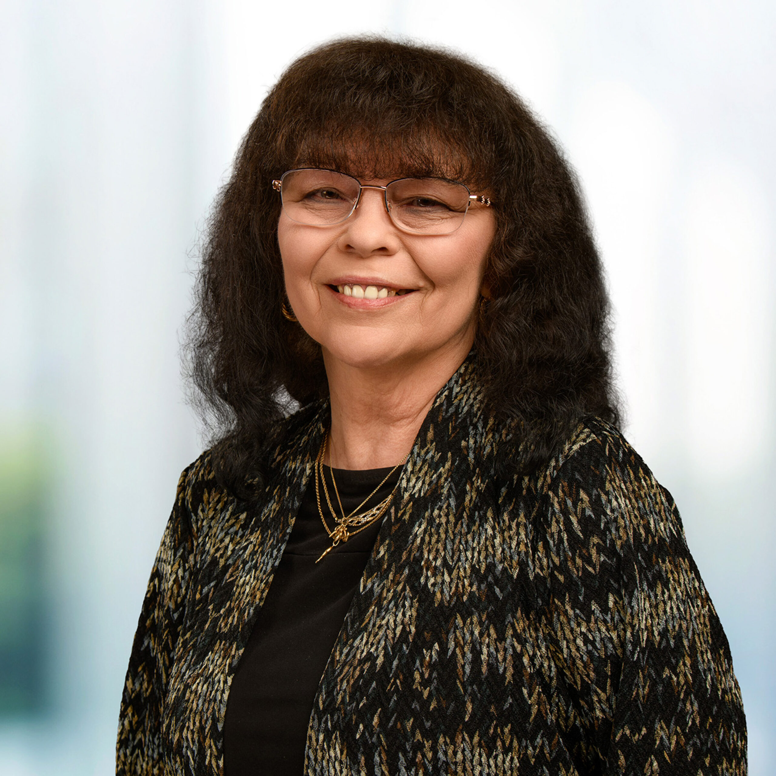 Smiling older woman with shoulder-length dark wavy hair, glasses, a patterned black-and-gold jacket, and layered gold necklaces against a soft blurred background.