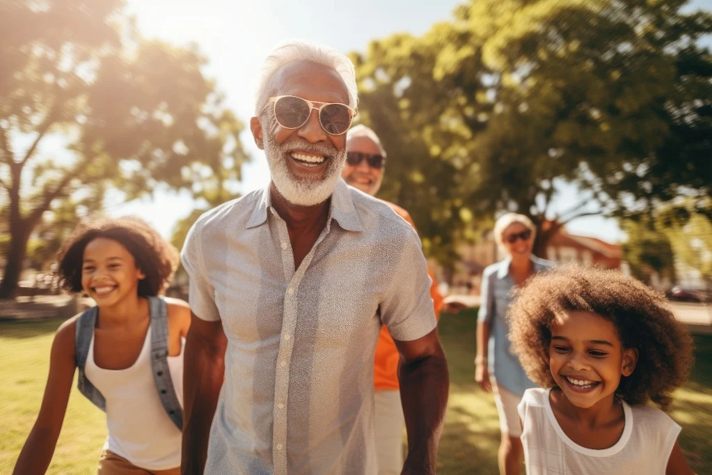 Smiling older man with white hair and beard wearing sunglasses leads a laughing multi-generation family, including two girls with curly hair, walking together in a sunlit park with trees in the background.