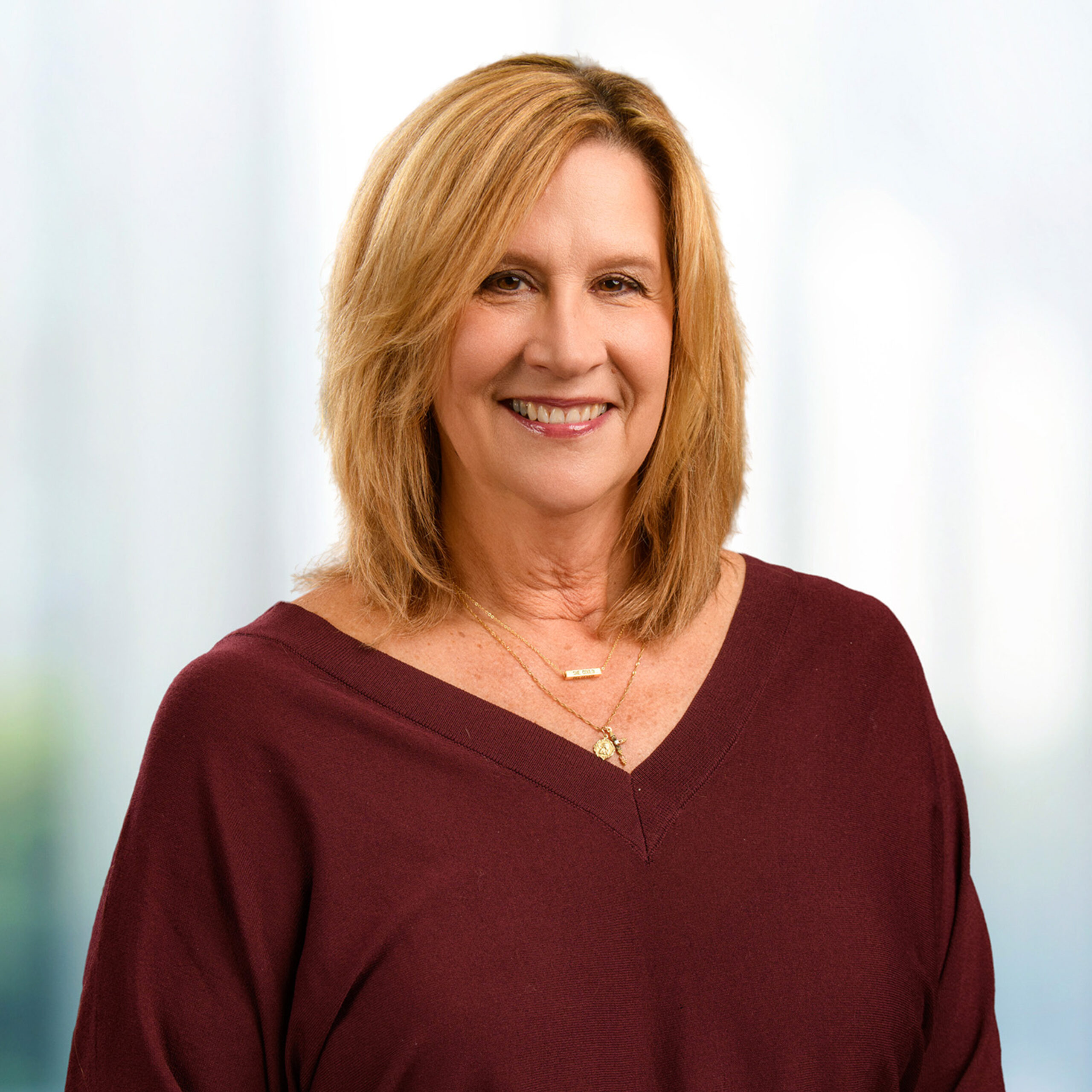 Smiling woman with shoulder-length blonde hair in a maroon V-neck and gold necklaces, soft blurred background.