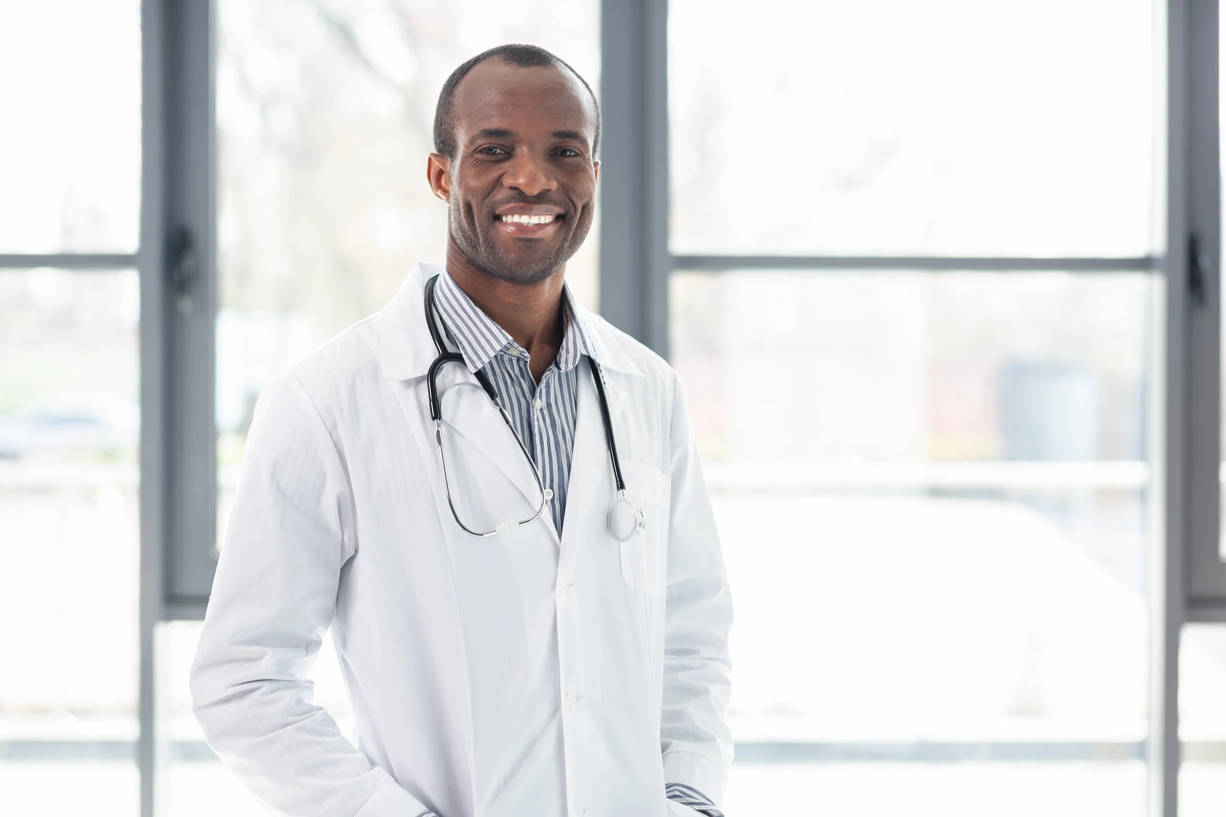 Smiling male doctor wearing a white lab coat and stethoscope over a striped shirt, standing in front of large bright windows.