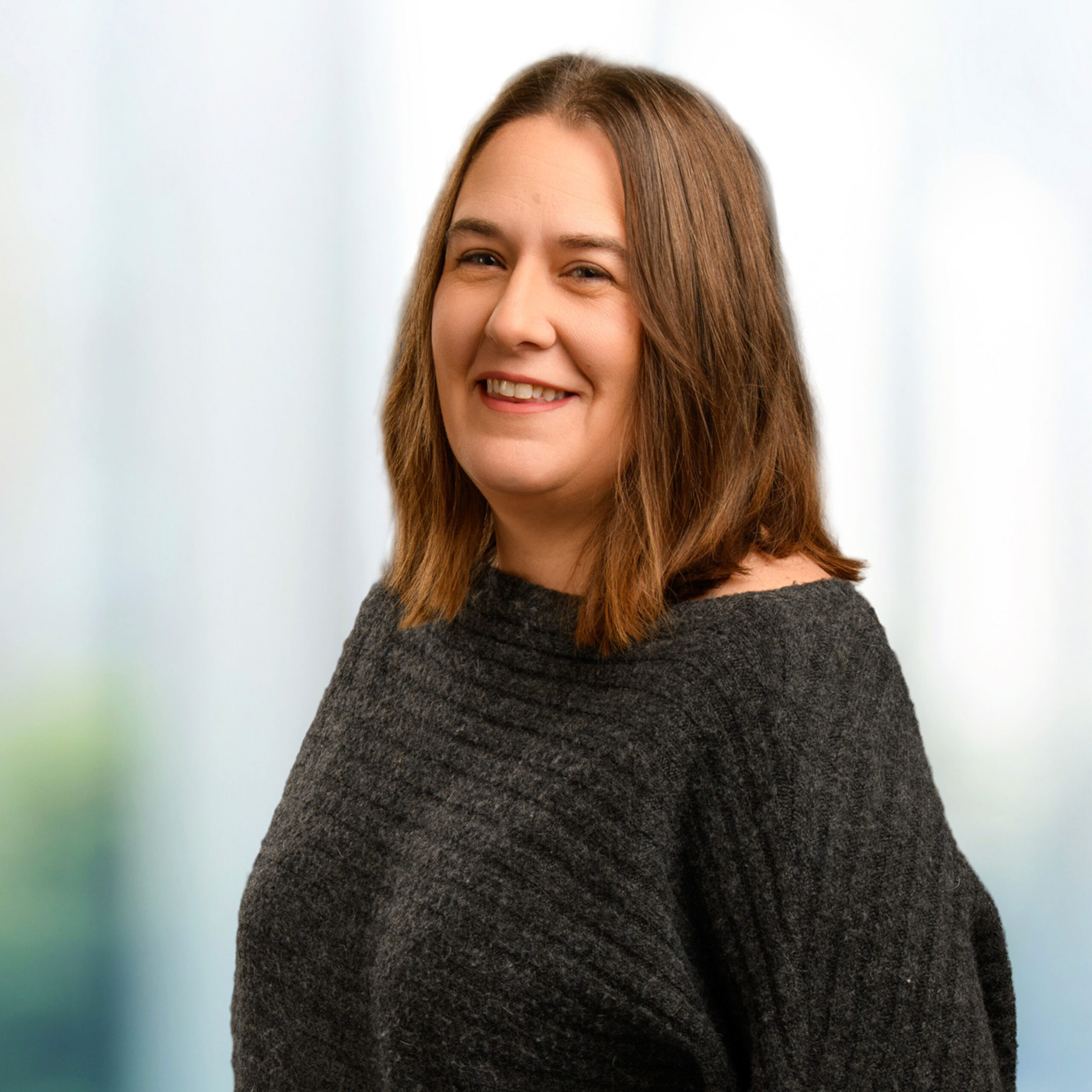 Smiling woman with shoulder-length brown hair wearing a dark gray textured sweater against a soft, blurred light background.
