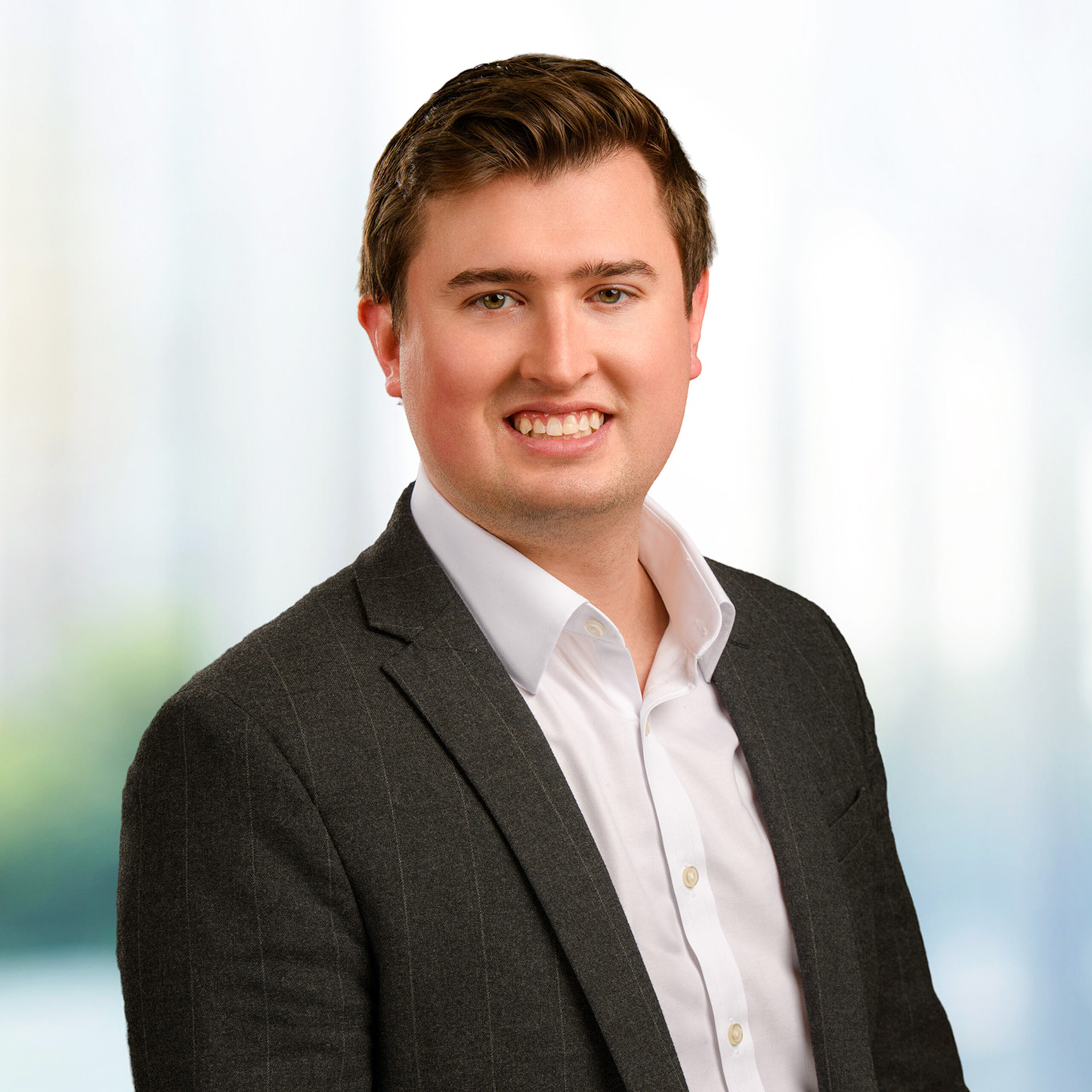 Smiling young man in dark pinstripe blazer and white dress shirt against a soft, light office-like background.