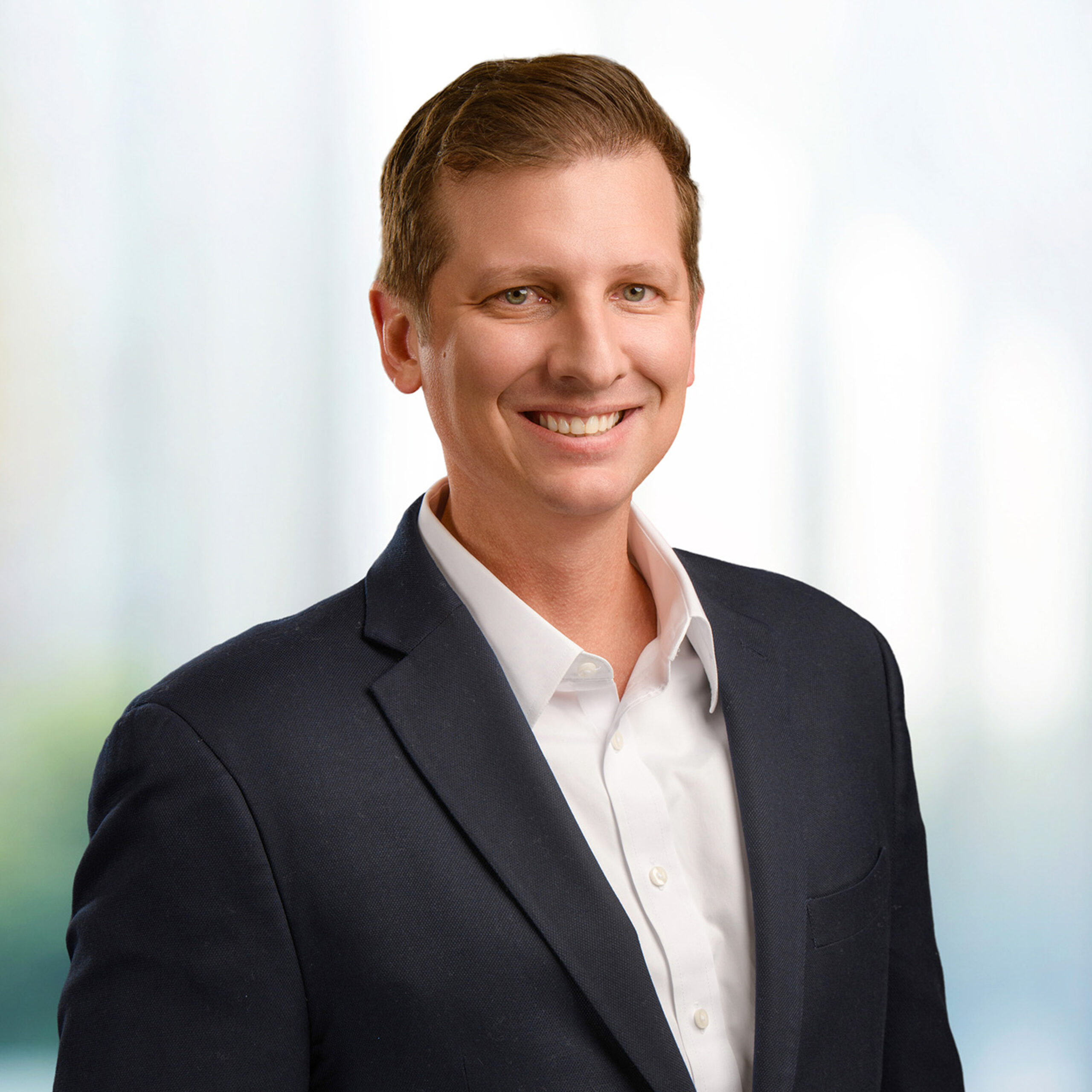 Smiling man with short brown hair wearing a navy blazer and white shirt in a bright professional headshot.
