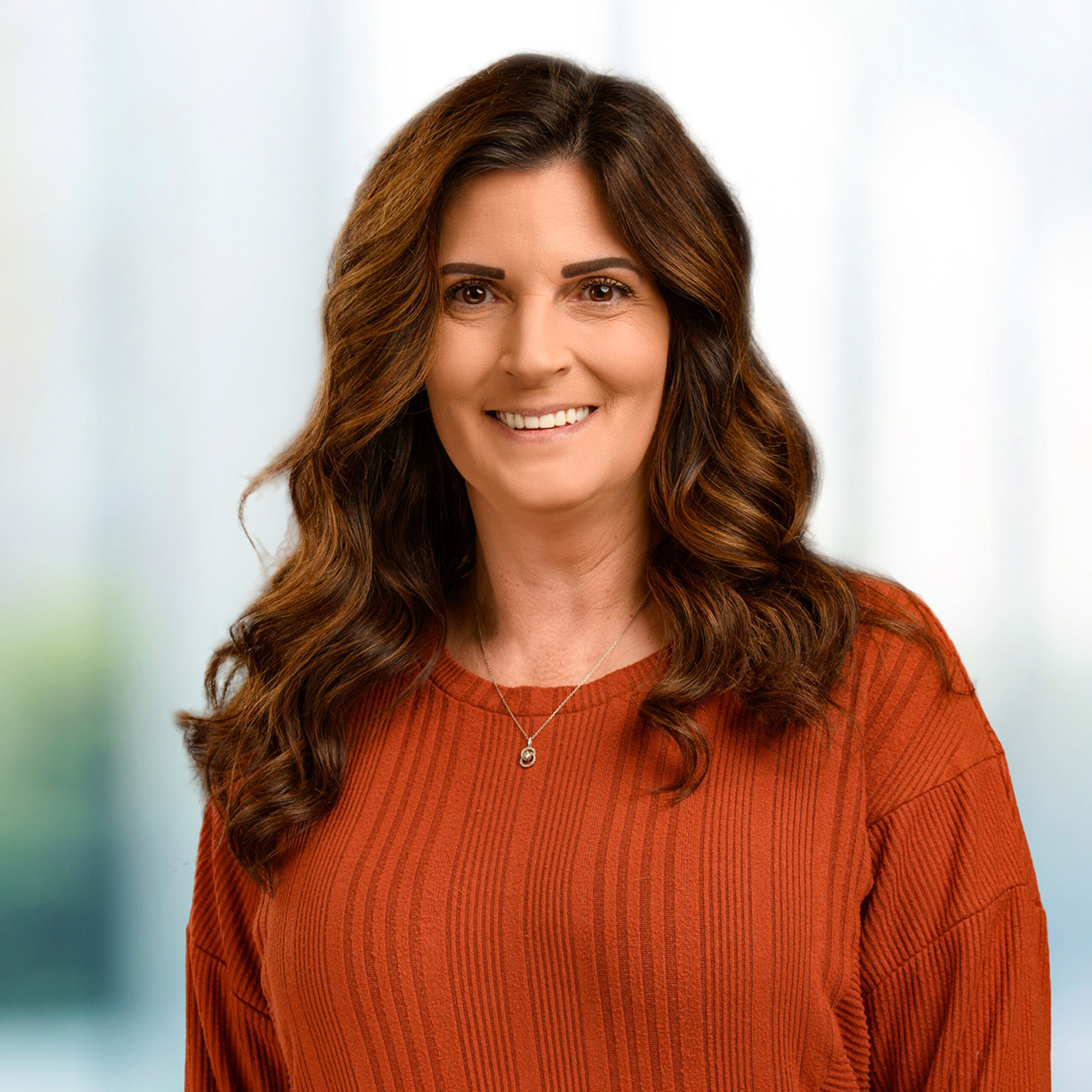 Smiling woman with wavy brown hair in an orange ribbed sweater wearing a pendant necklace, blurred background.