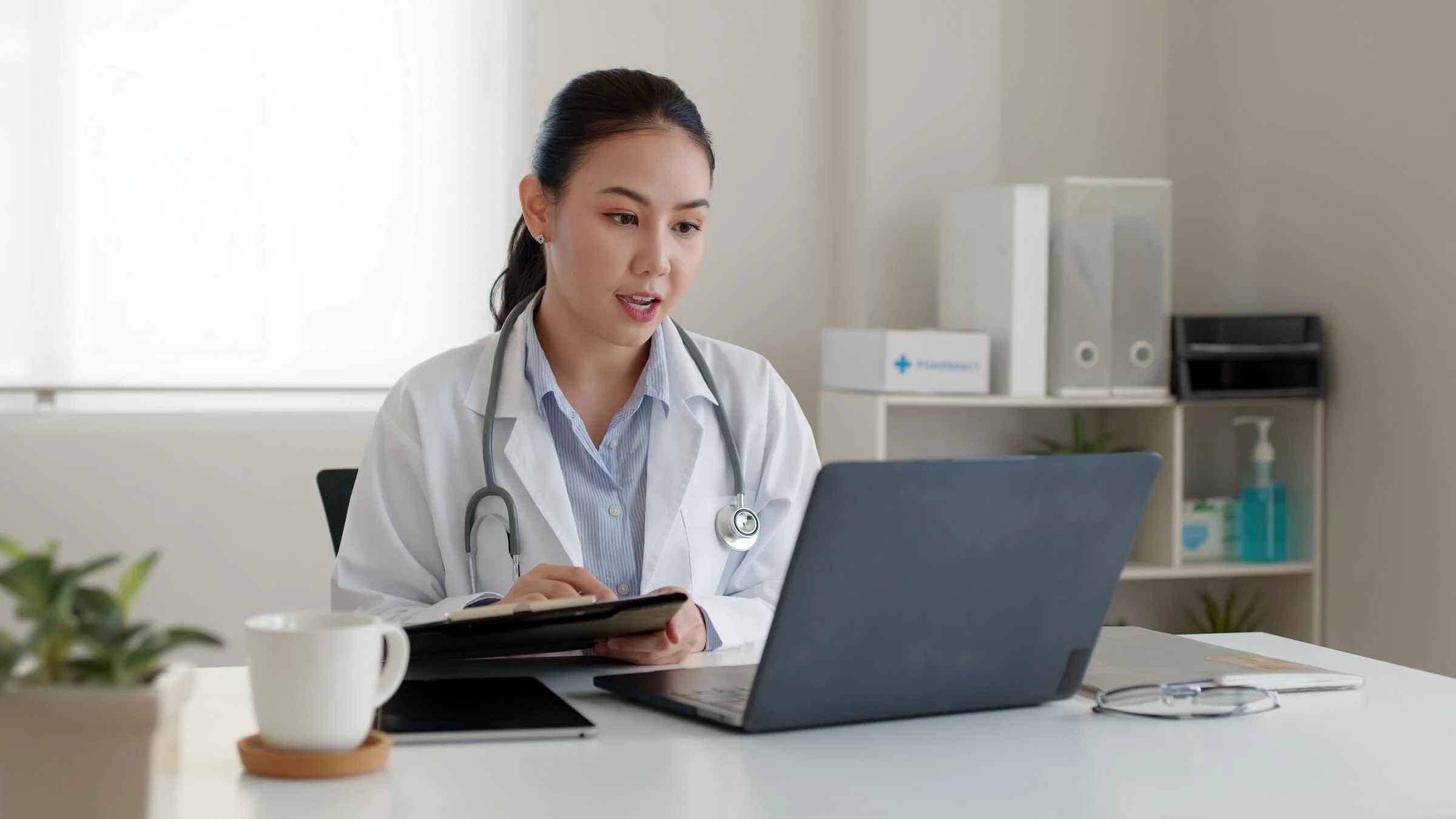 Female doctor in a white coat with a stethoscope around her neck sits at a desk holding a clipboard and speaking to a laptop in a bright office with shelves, a coffee mug, and a small potted plant.