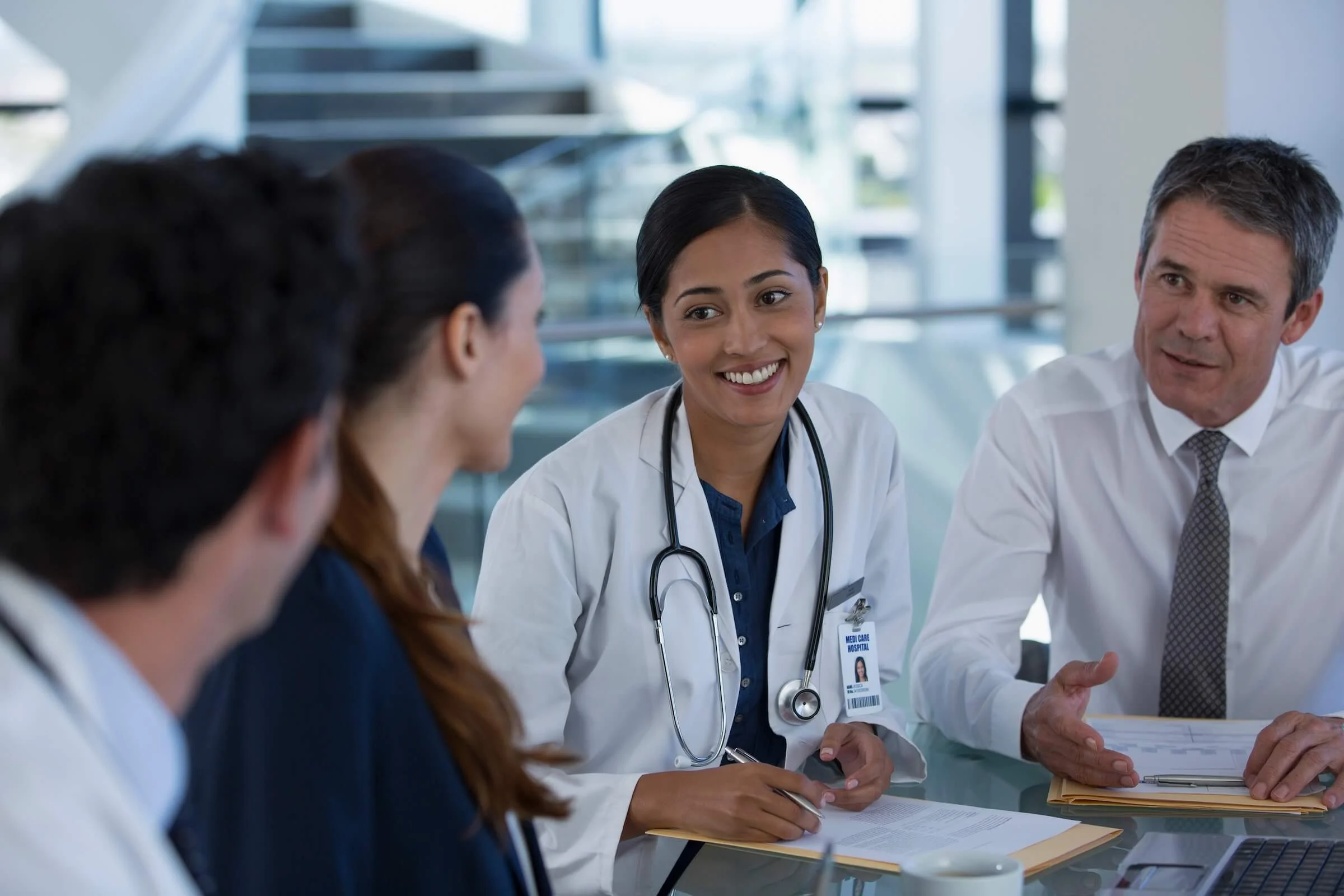 A smiling doctor in a white coat with a stethoscope and ID badge reviews paperwork and talks with colleagues around a conference table as a male colleague gestures.