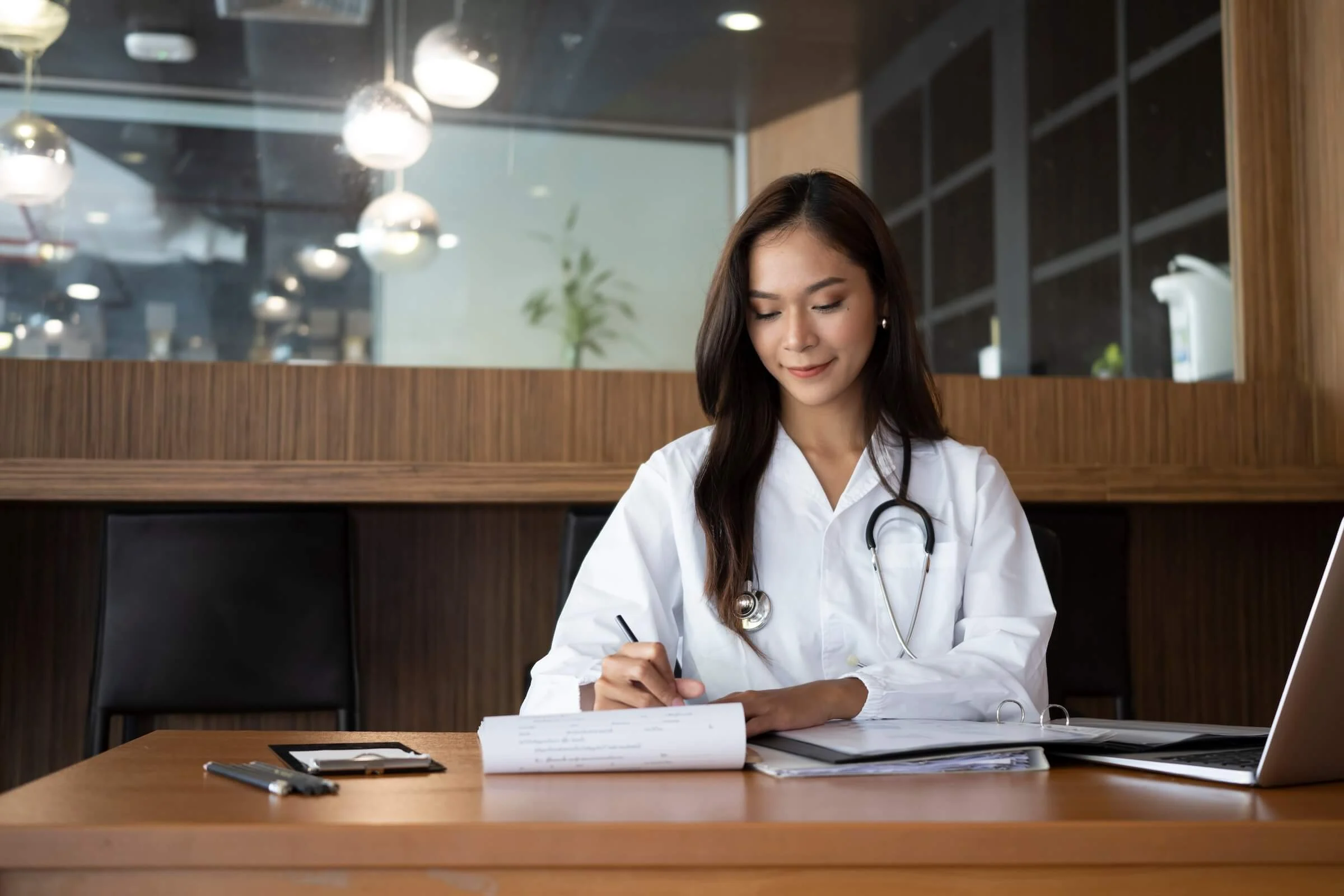 Young female doctor in a white coat with a stethoscope around her neck sits at a wooden desk in a modern office, smiling slightly as she writes on medical forms with a laptop and clipboard nearby.