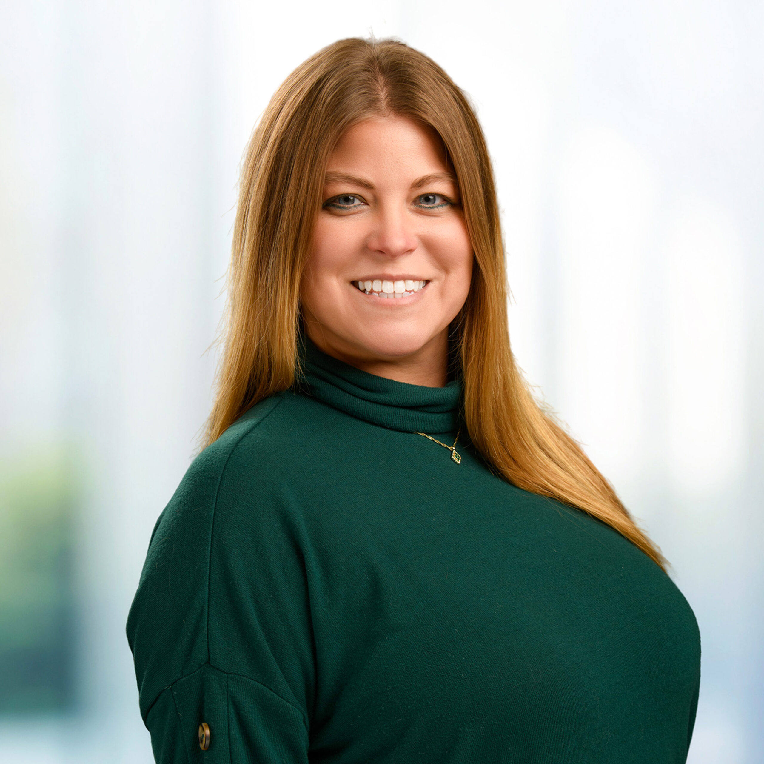 Smiling woman with long light brown hair wearing a dark green turtleneck and gold necklace against a blurred background.