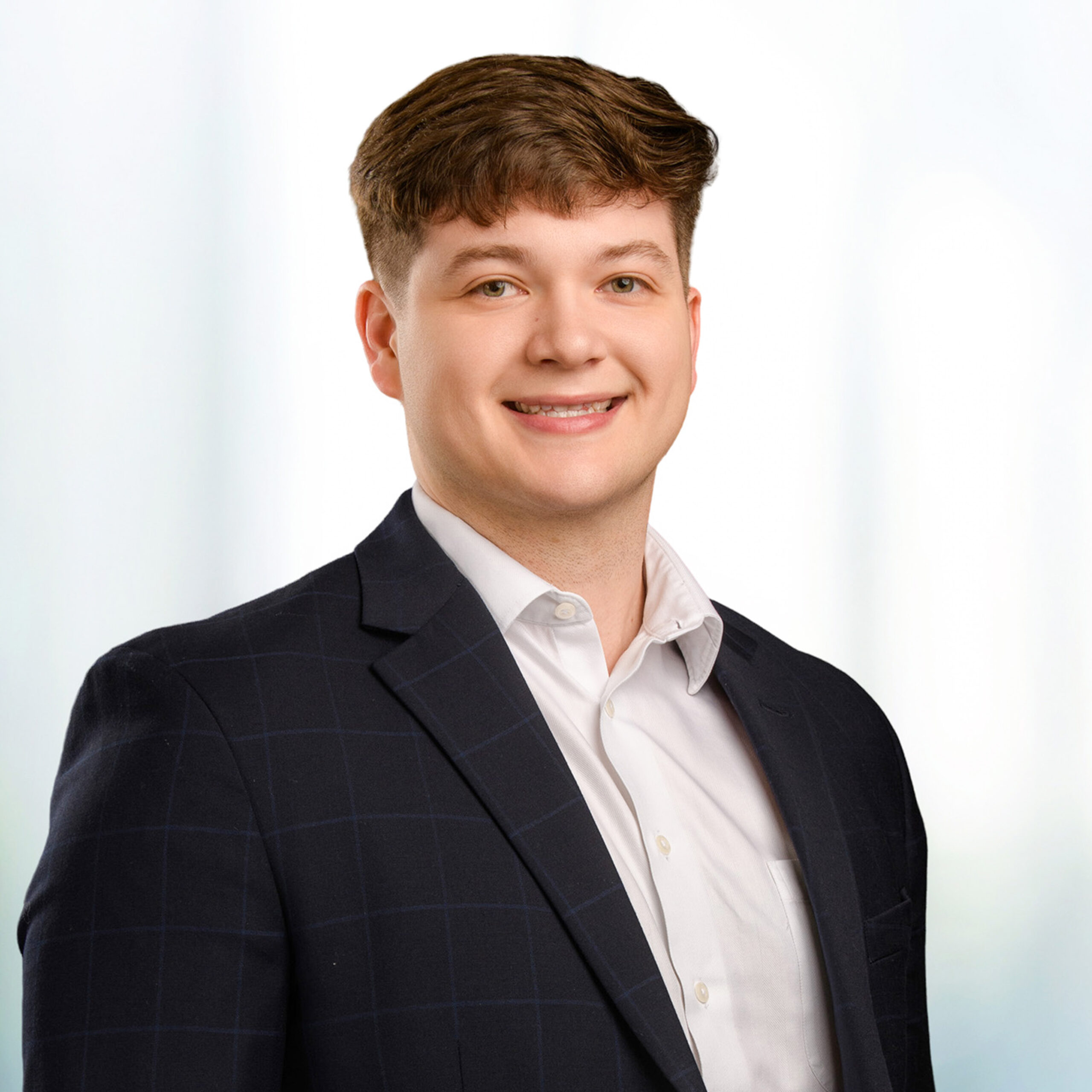 Young man with brown hair smiling, wearing a dark checked suit jacket and white dress shirt against a blurred background.