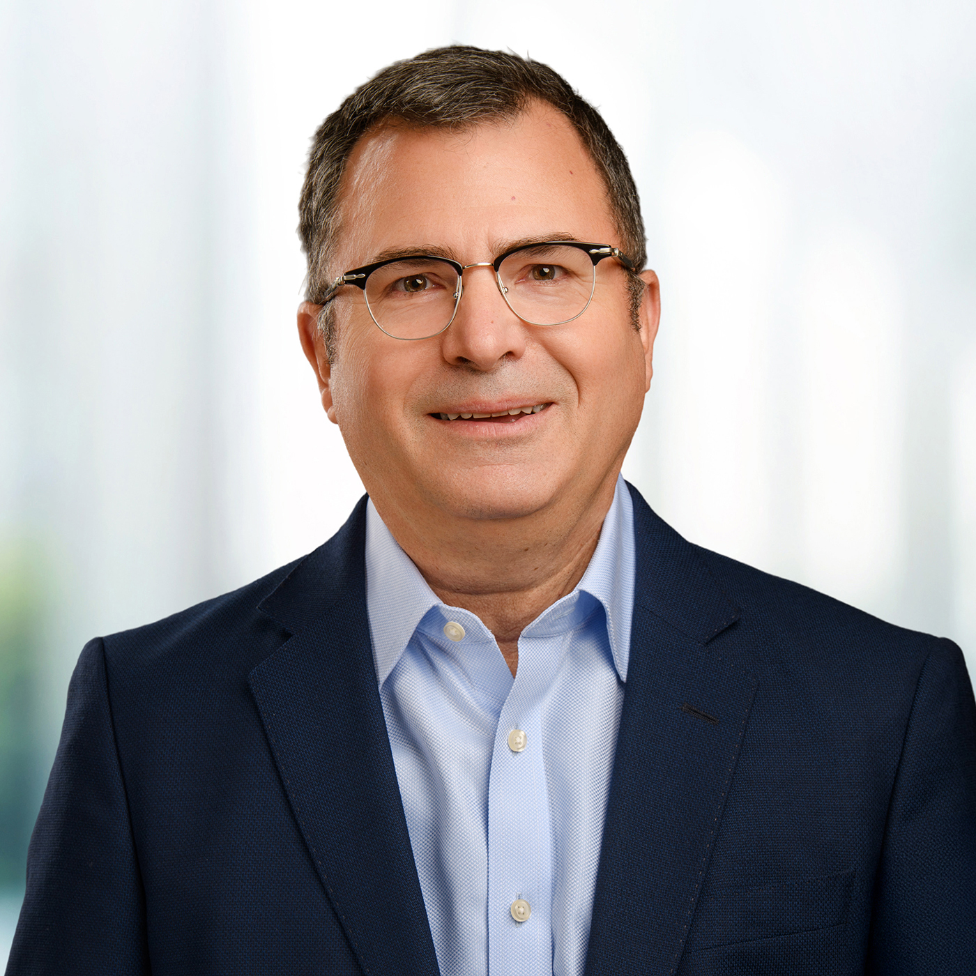 Man with glasses wearing a navy blazer and light blue shirt, smiling against a soft blurred background.