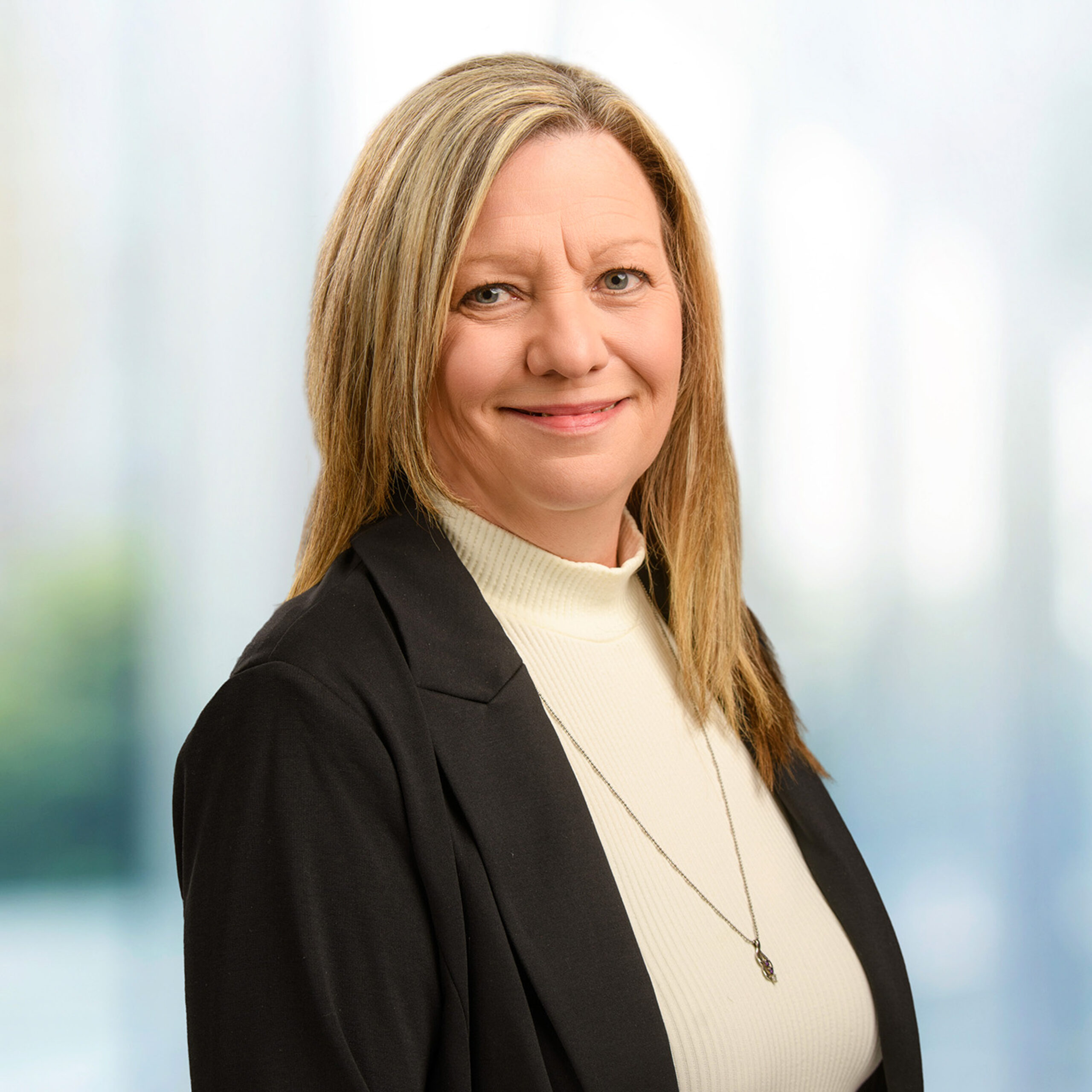 Woman with shoulder-length blonde hair wearing a white turtleneck, black blazer and pendant necklace, smiling.