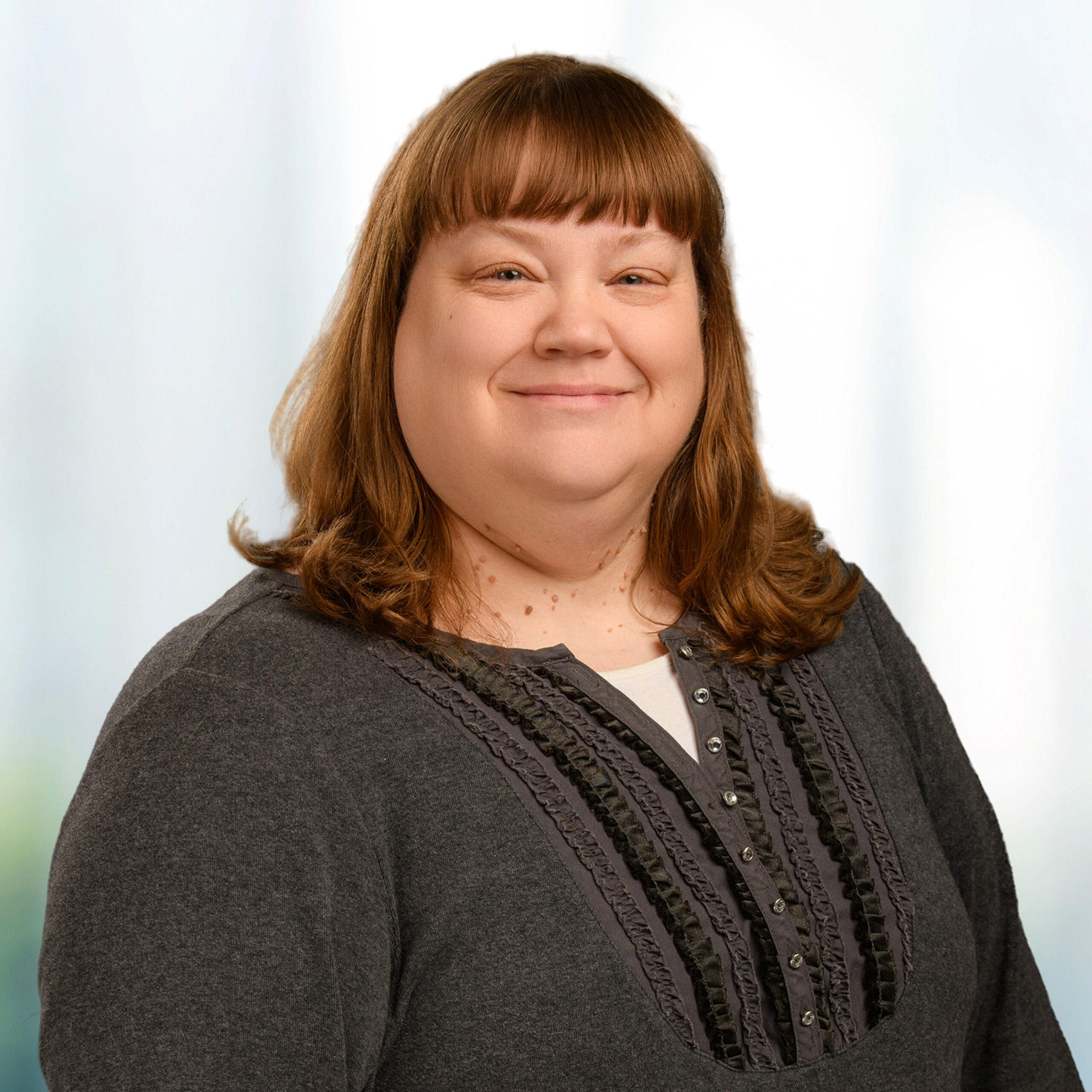 Smiling woman with shoulder-length brown hair and bangs wearing a dark gray blouse with ruffled trim on a light background.
