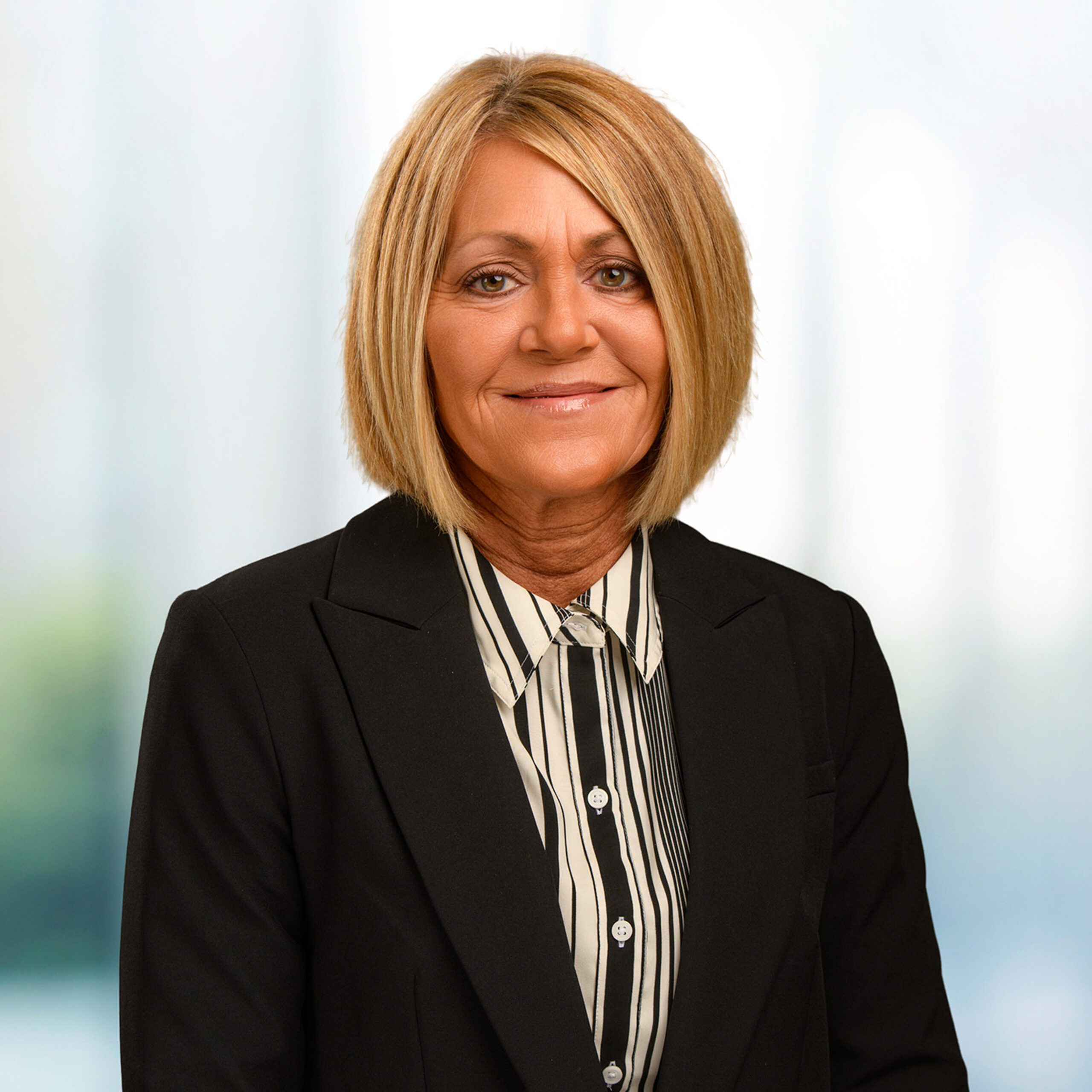 Smiling woman with chin-length blonde bob wearing a black blazer and striped blouse against a soft office background.