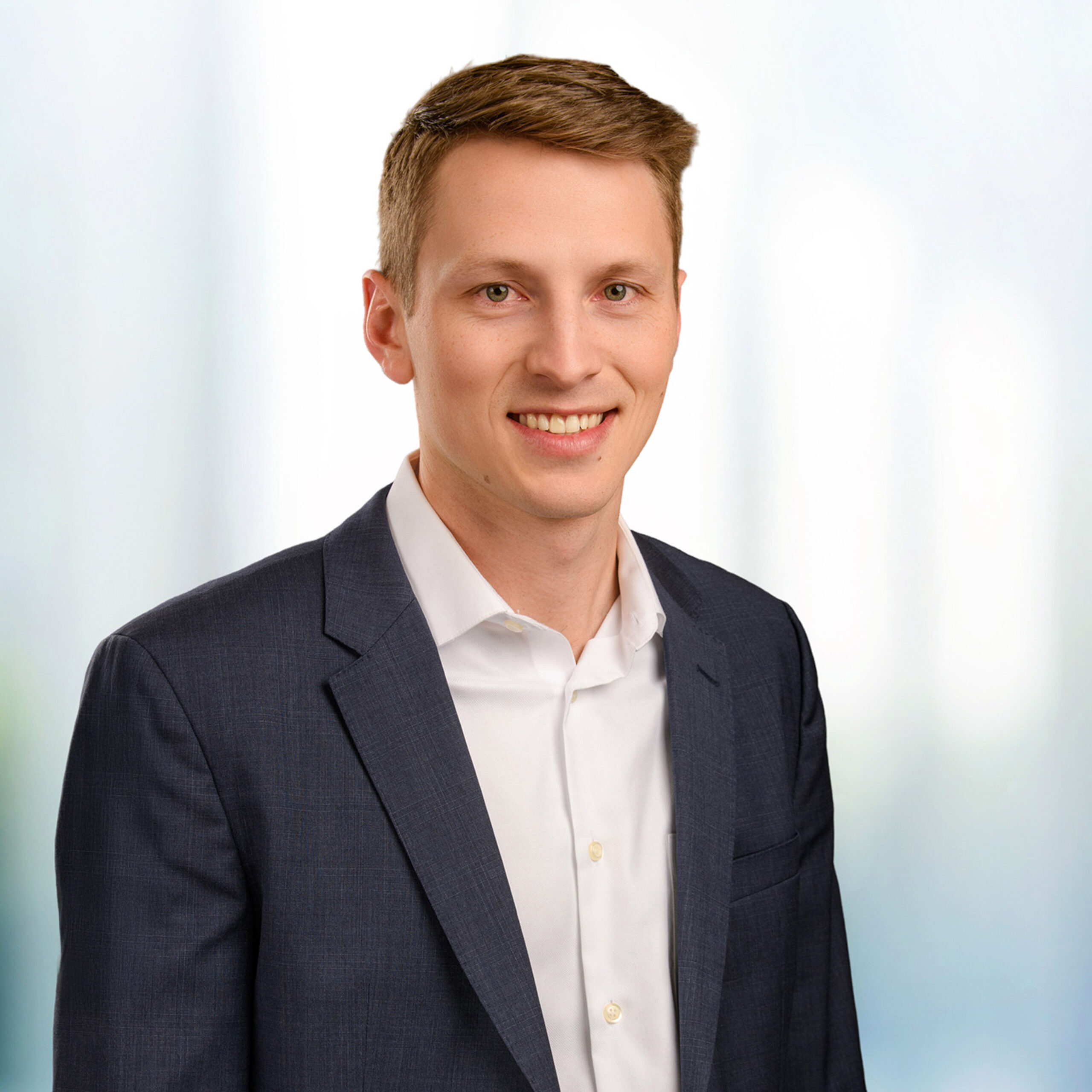 Young smiling man with short light brown hair wearing a dark suit jacket and white shirt against a blurred office background.