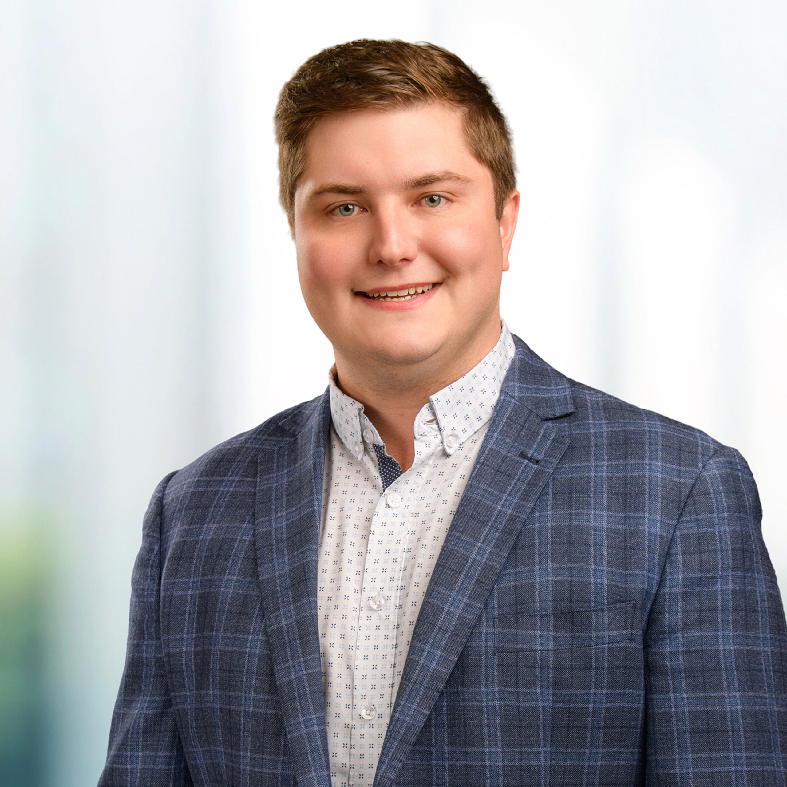 Young smiling man wearing a blue plaid blazer and patterned white button‑up shirt against a soft blurred background.