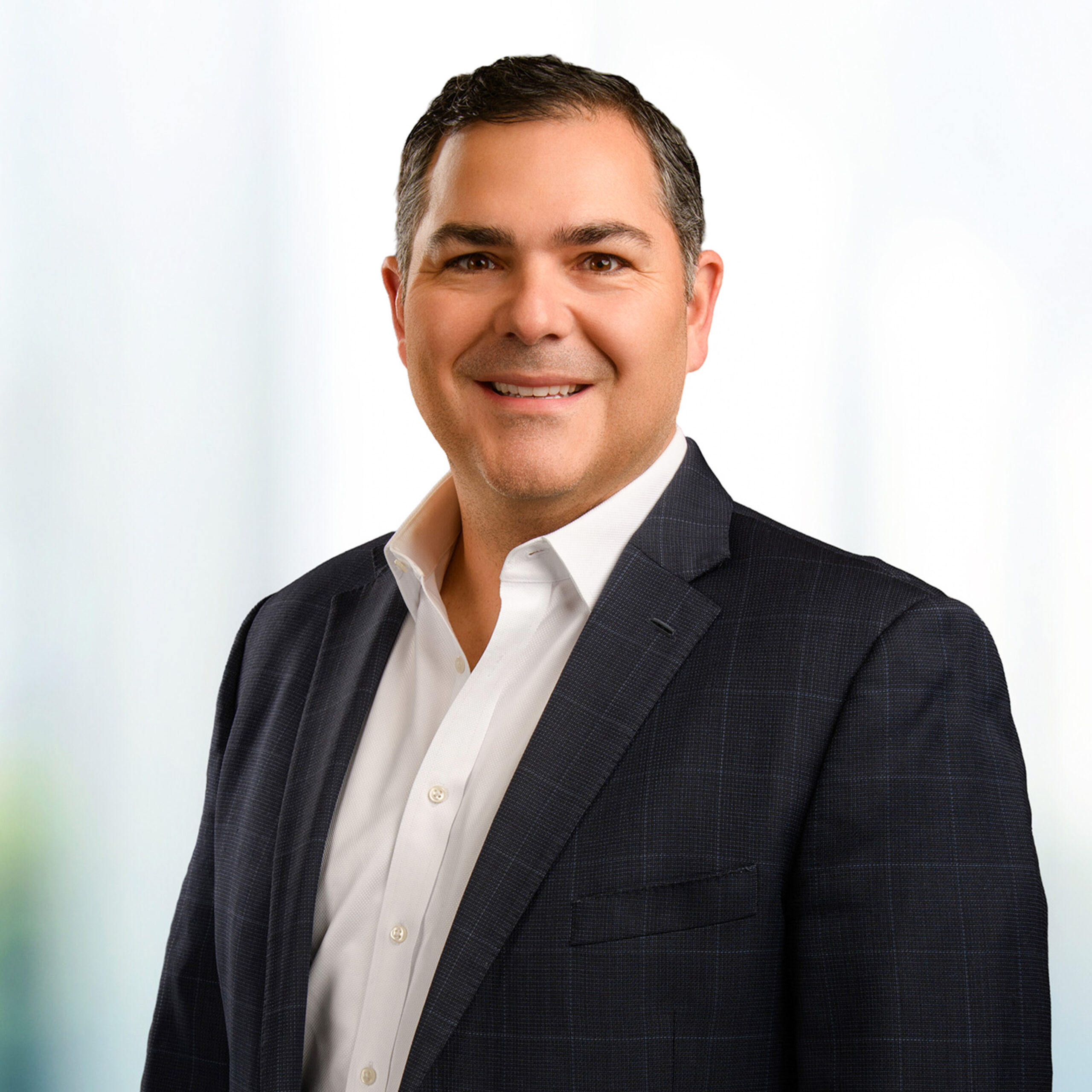 Smiling man wearing a dark checked suit jacket and white dress shirt against a soft, light background.