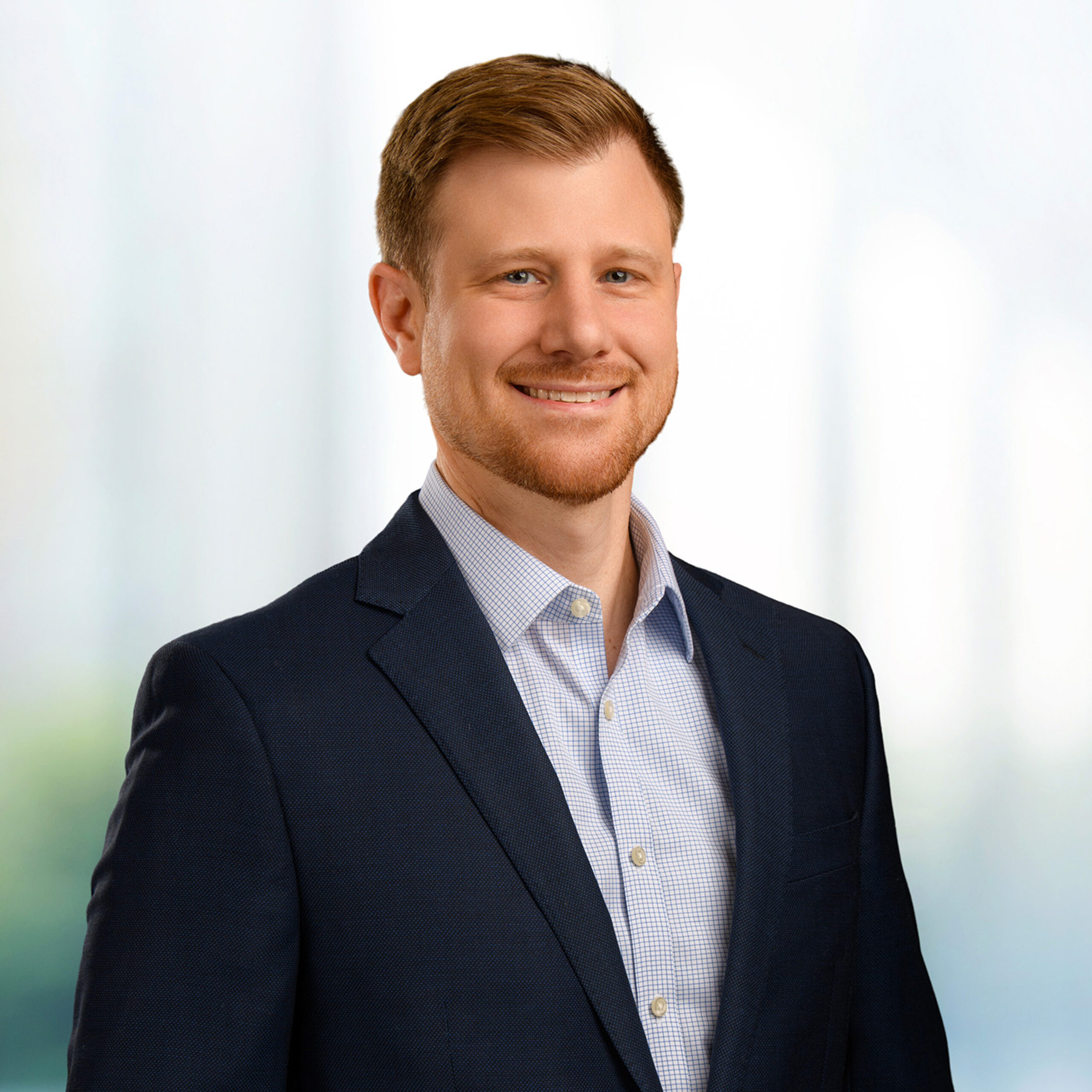Smiling man with short light brown hair and trimmed beard wearing a navy blazer and checked shirt on a blurred background.