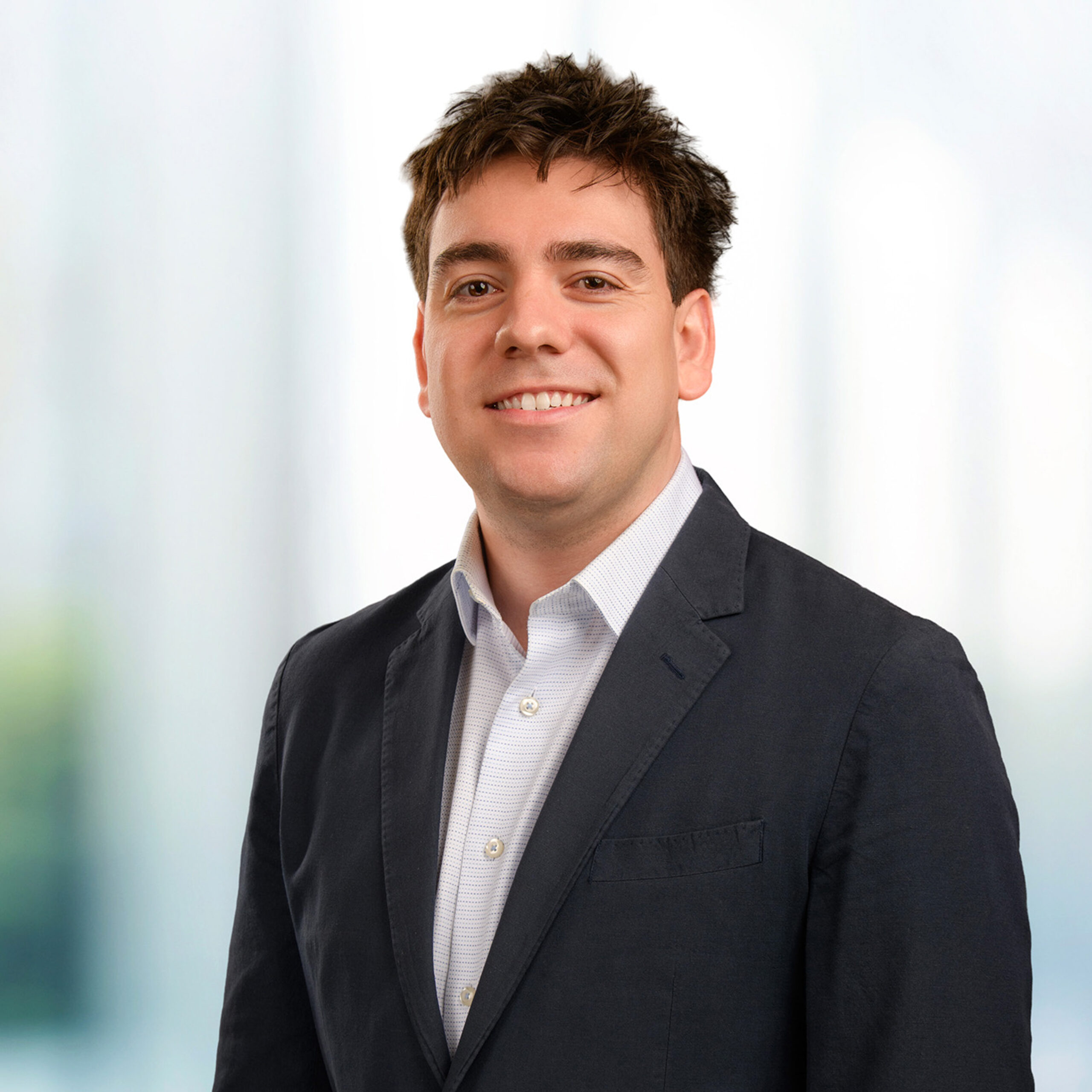 Smiling young man with short dark hair wearing a dark blazer and white shirt against a soft, blurred light background.
