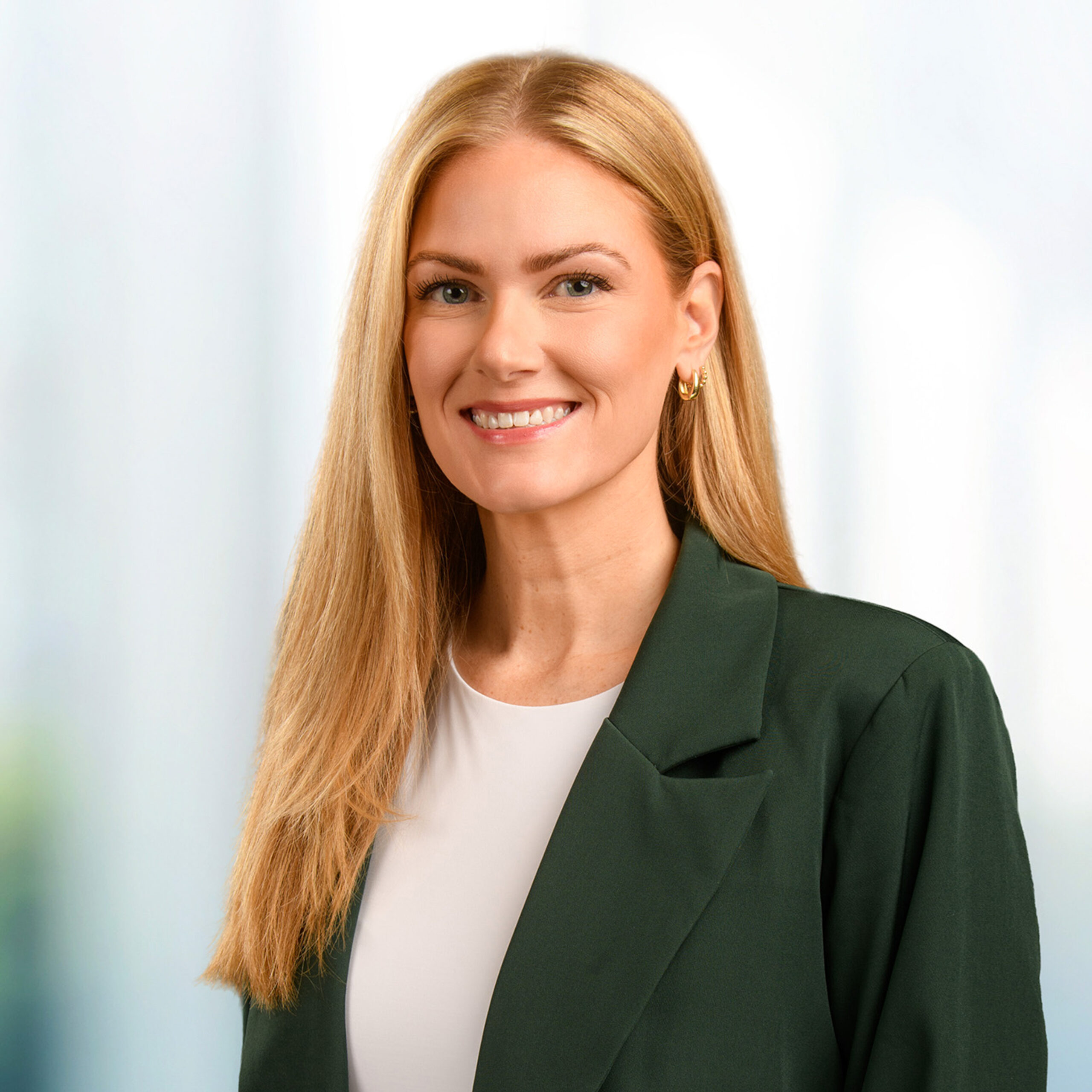 Smiling blonde woman in a dark green blazer and white top wearing gold hoop earrings before a soft blurred background.