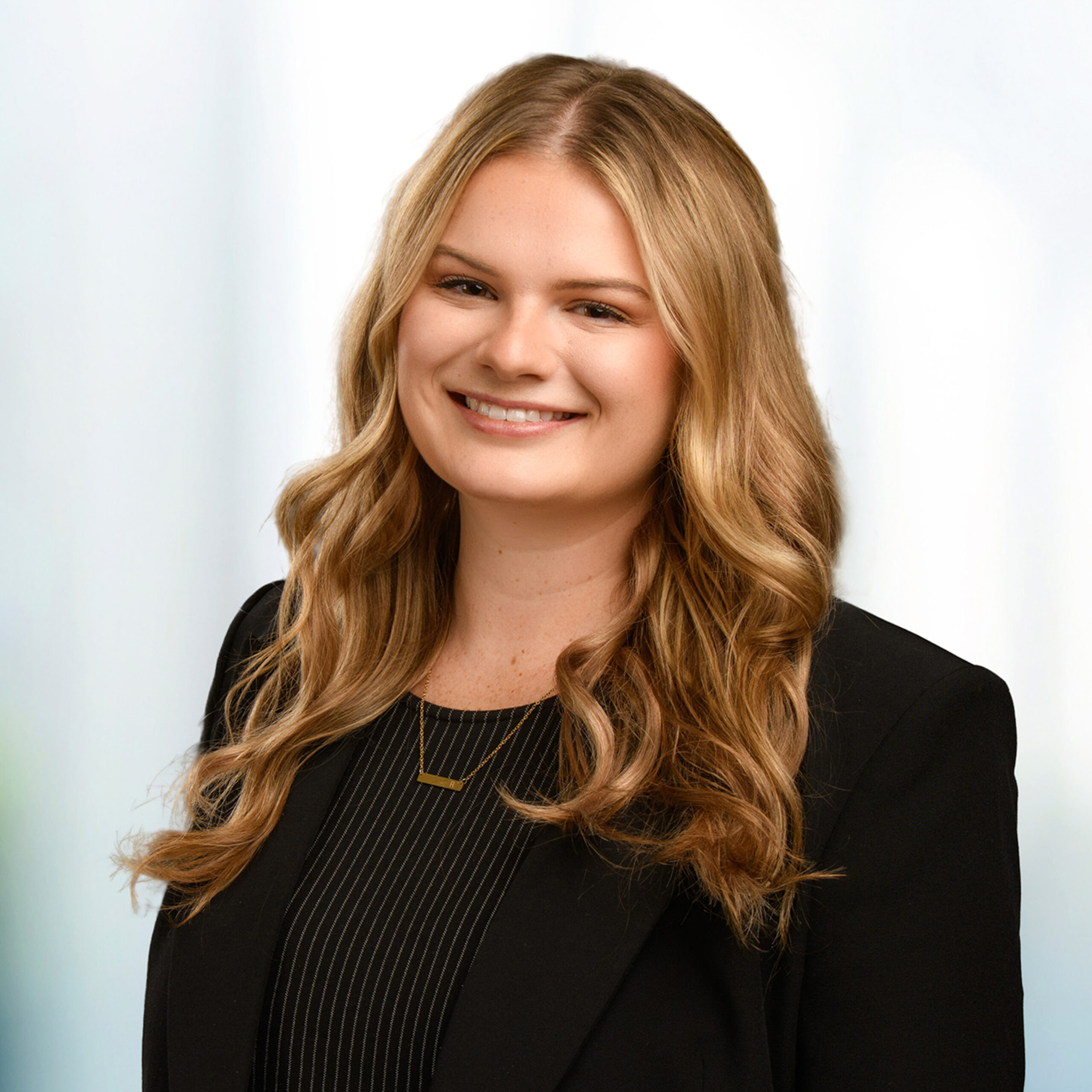 Young woman with wavy blonde hair smiling, wearing a black blazer and gold bar necklace against a soft light background.