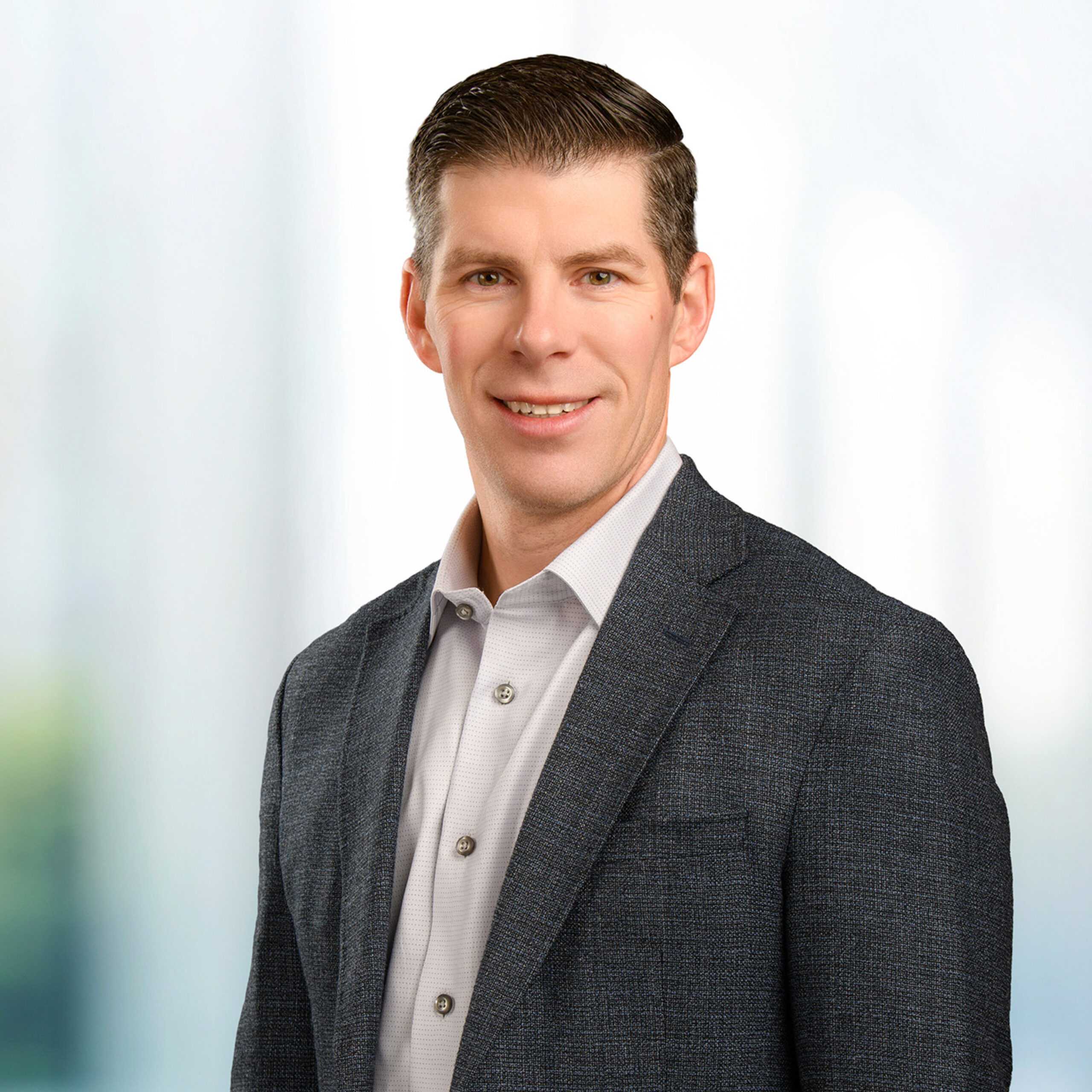 Smiling man in a dark gray blazer and light dress shirt with neatly combed short hair against a soft blurred background.