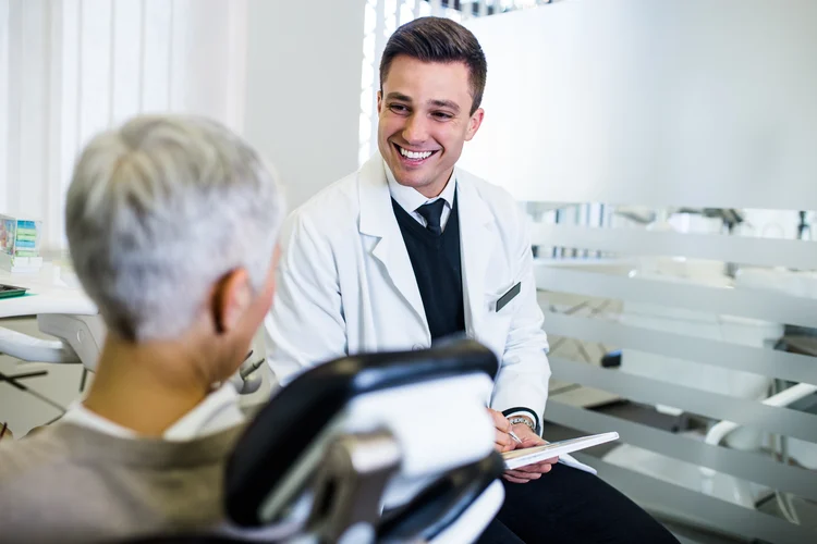 Smiling male dentist in a white coat holding a tablet talks with an older patient seated in a dental chair in a bright clinic.