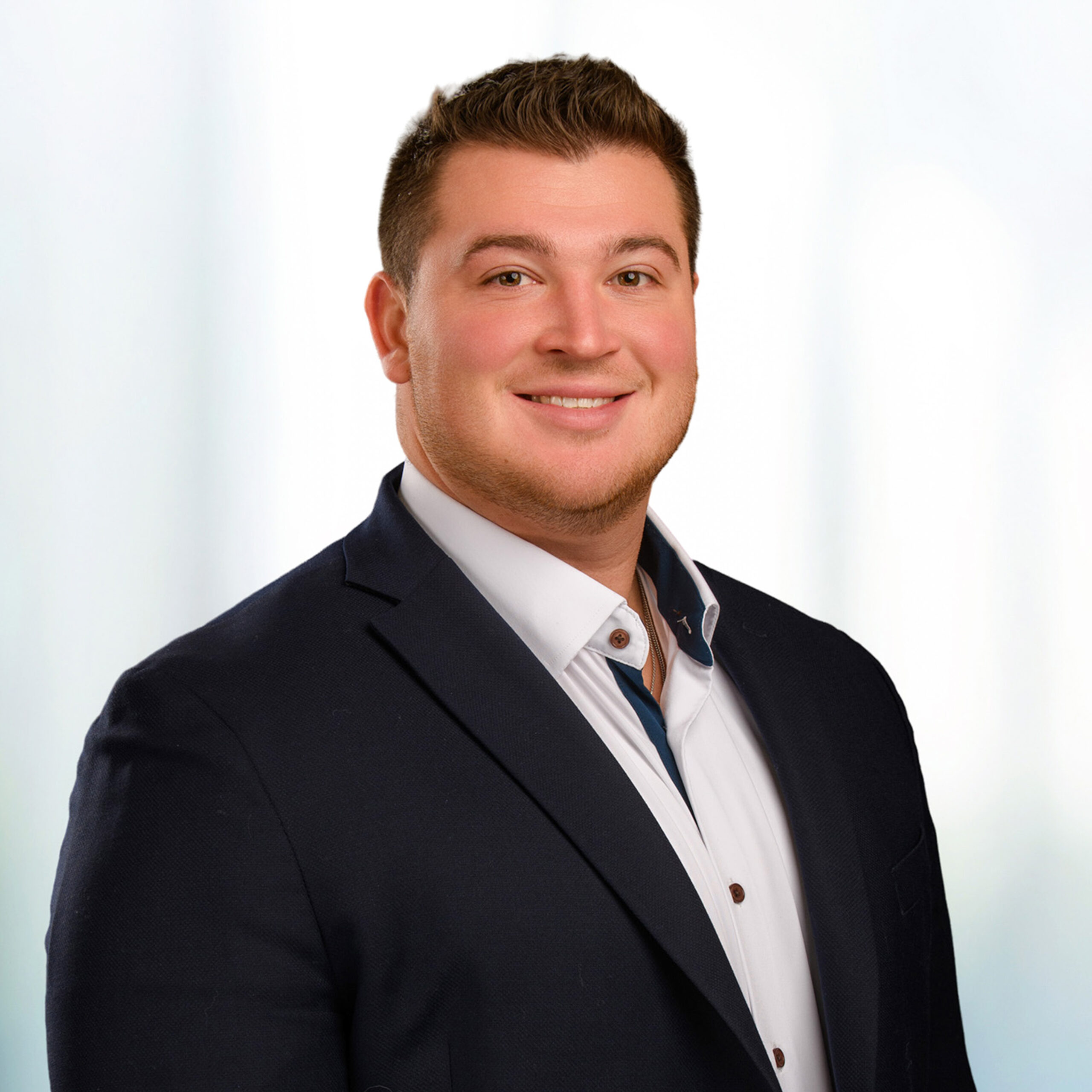 Smiling young man with short brown hair and light stubble, wearing a dark blazer and white shirt against a soft blurred background.