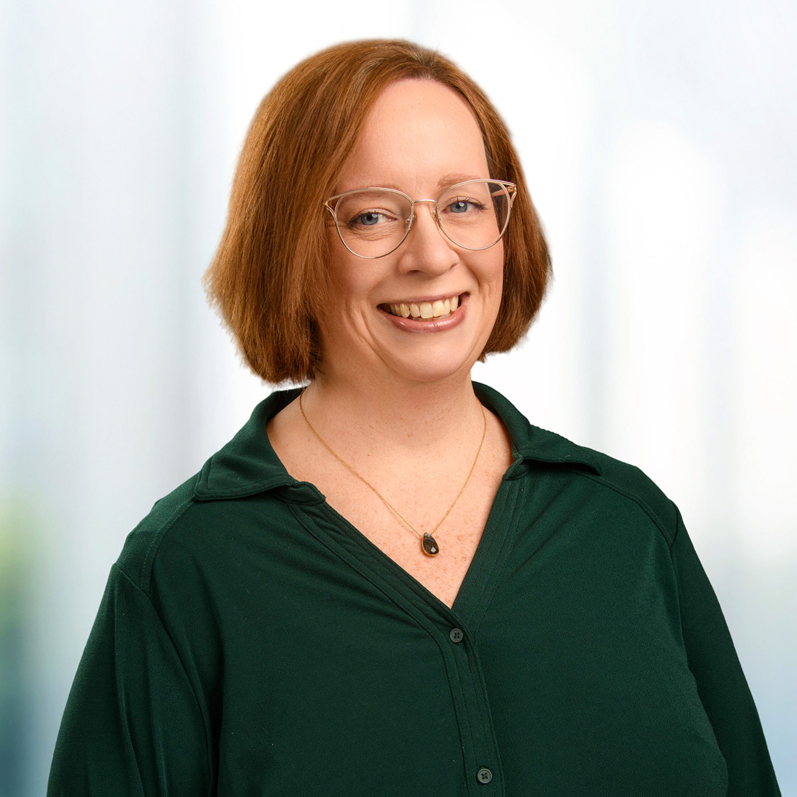 Smiling woman with auburn hair and glasses wearing a dark green blouse and pendant necklace on a blurred background.