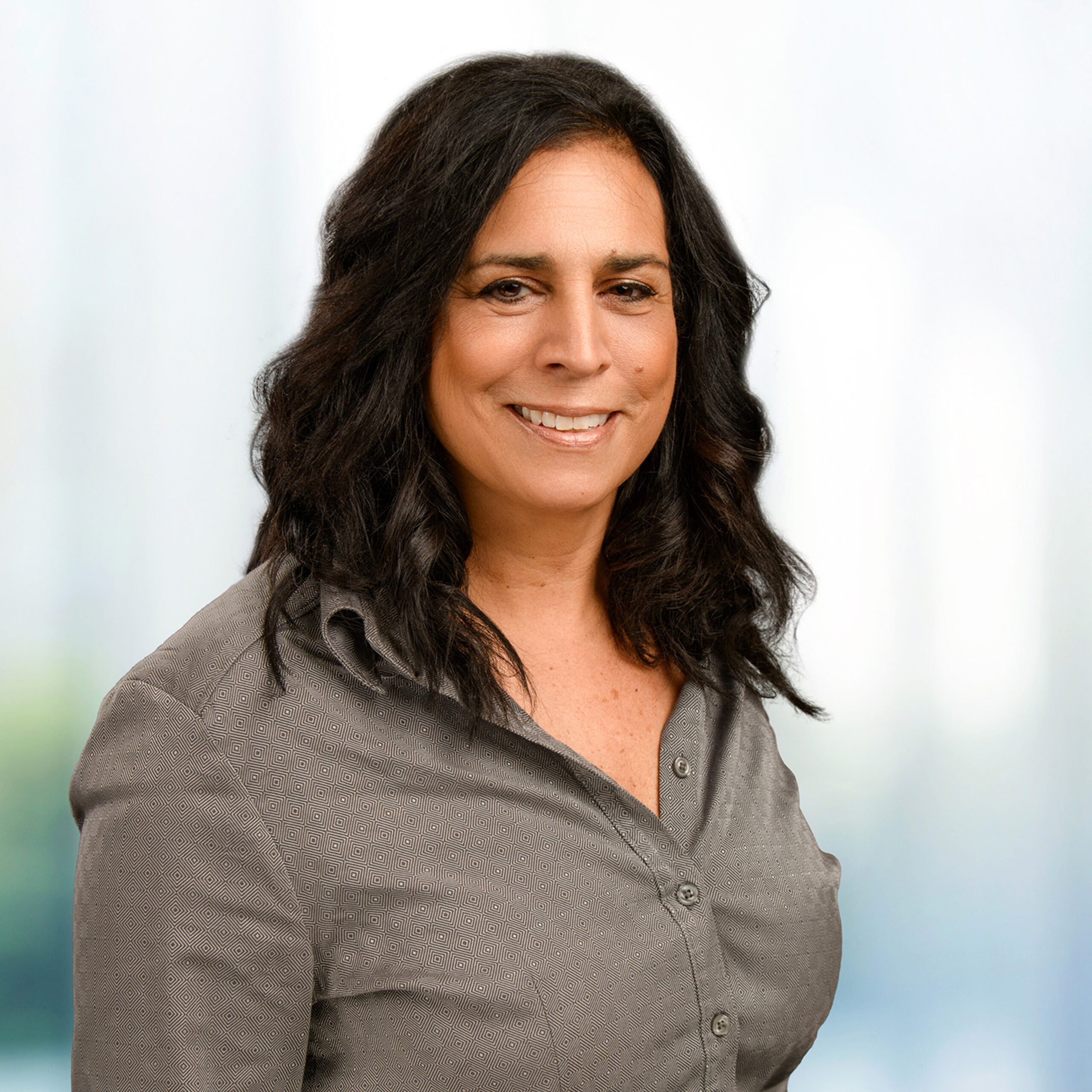 Smiling middle-aged woman with wavy dark hair wearing a gray patterned button-up shirt before a soft blurred background