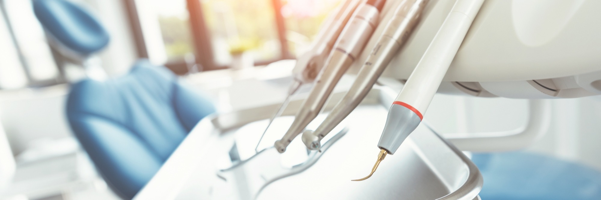 Close-up of dental handpieces and a white ultrasonic scaler with a gold tip hanging over a tray, with a blurred blue dental chair and sunlit window in the background.