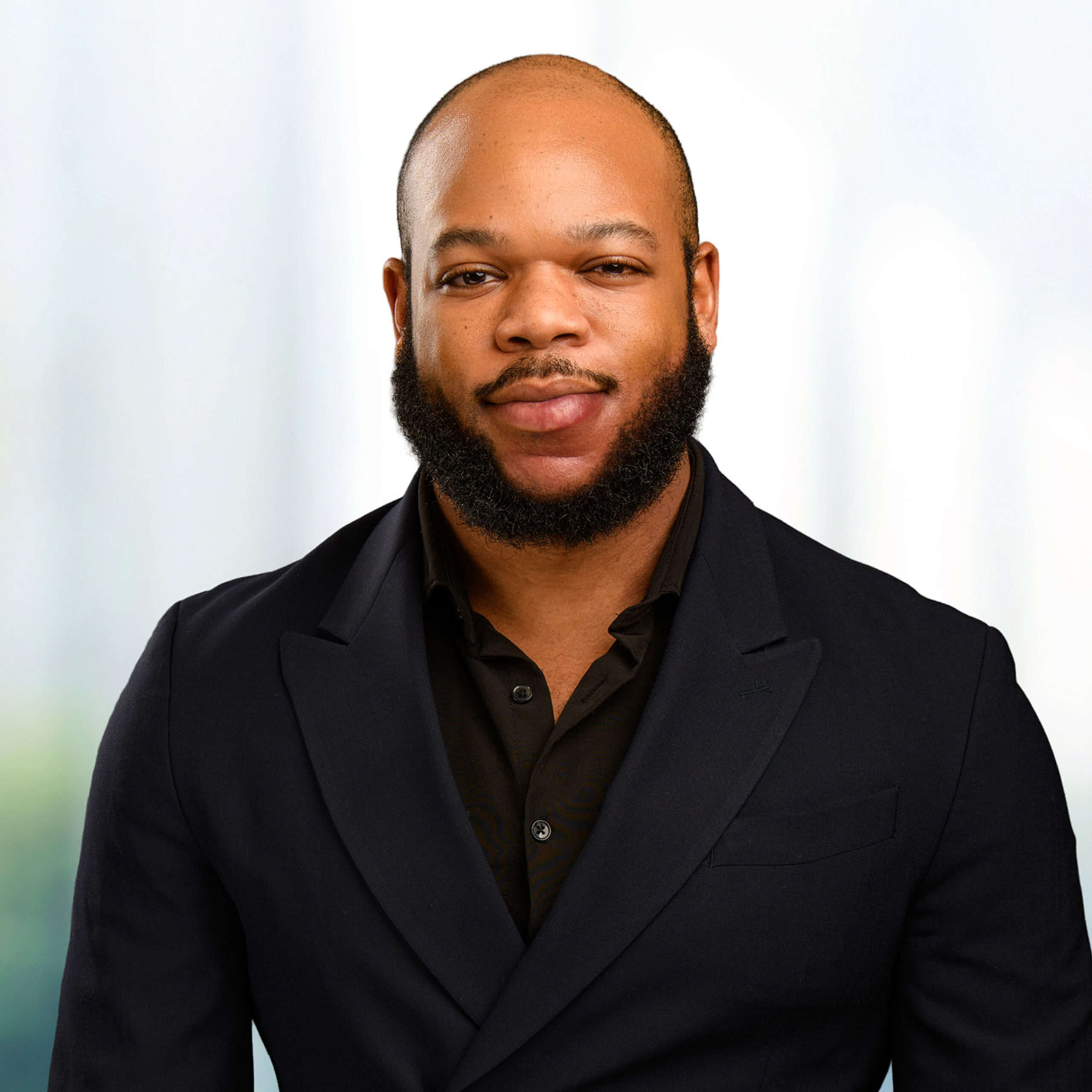 Headshot of a bald man with a trimmed beard wearing a black suit and shirt against a soft blurred background.