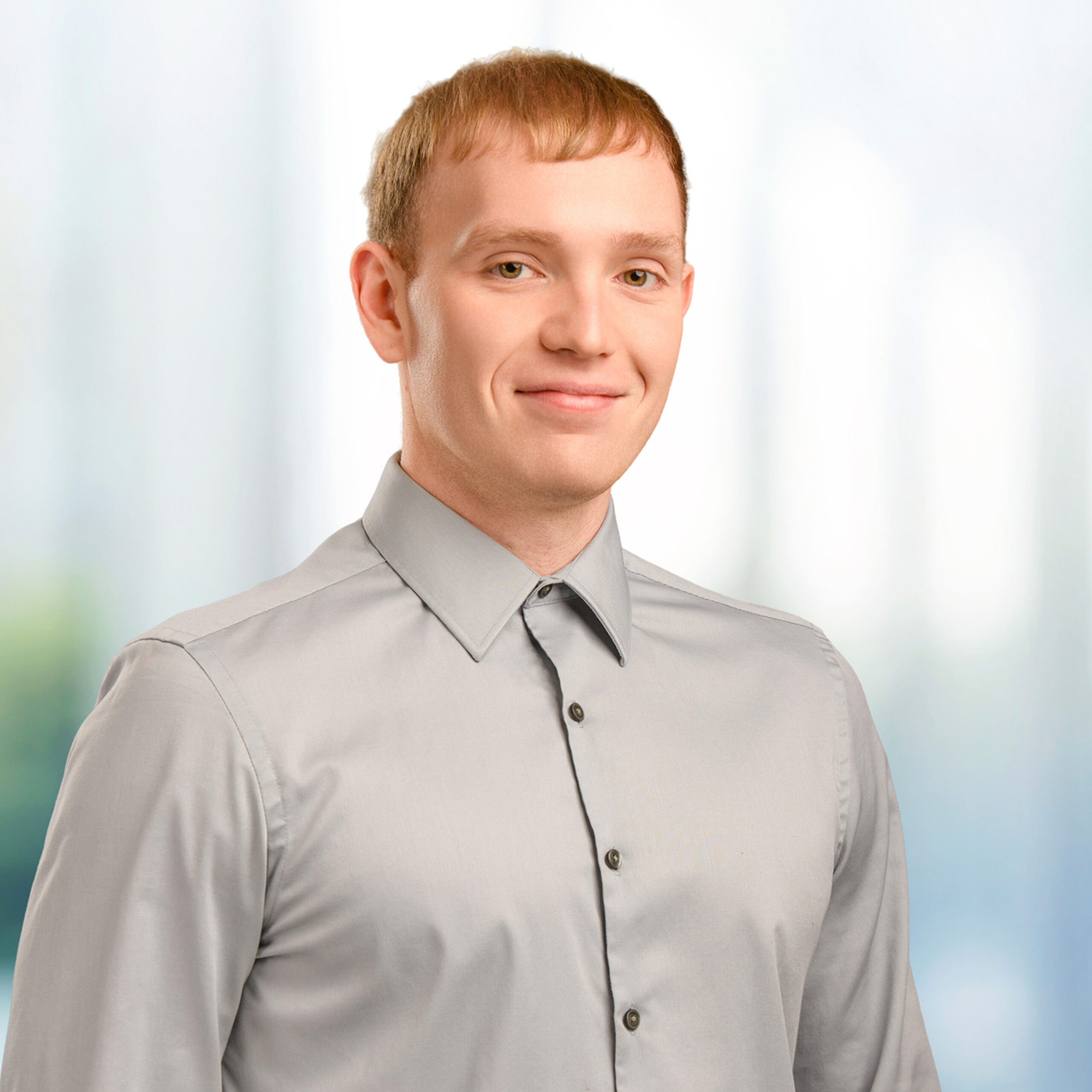 Man with short light brown hair and gray button-up shirt smiling slightly against a blurred bright background.