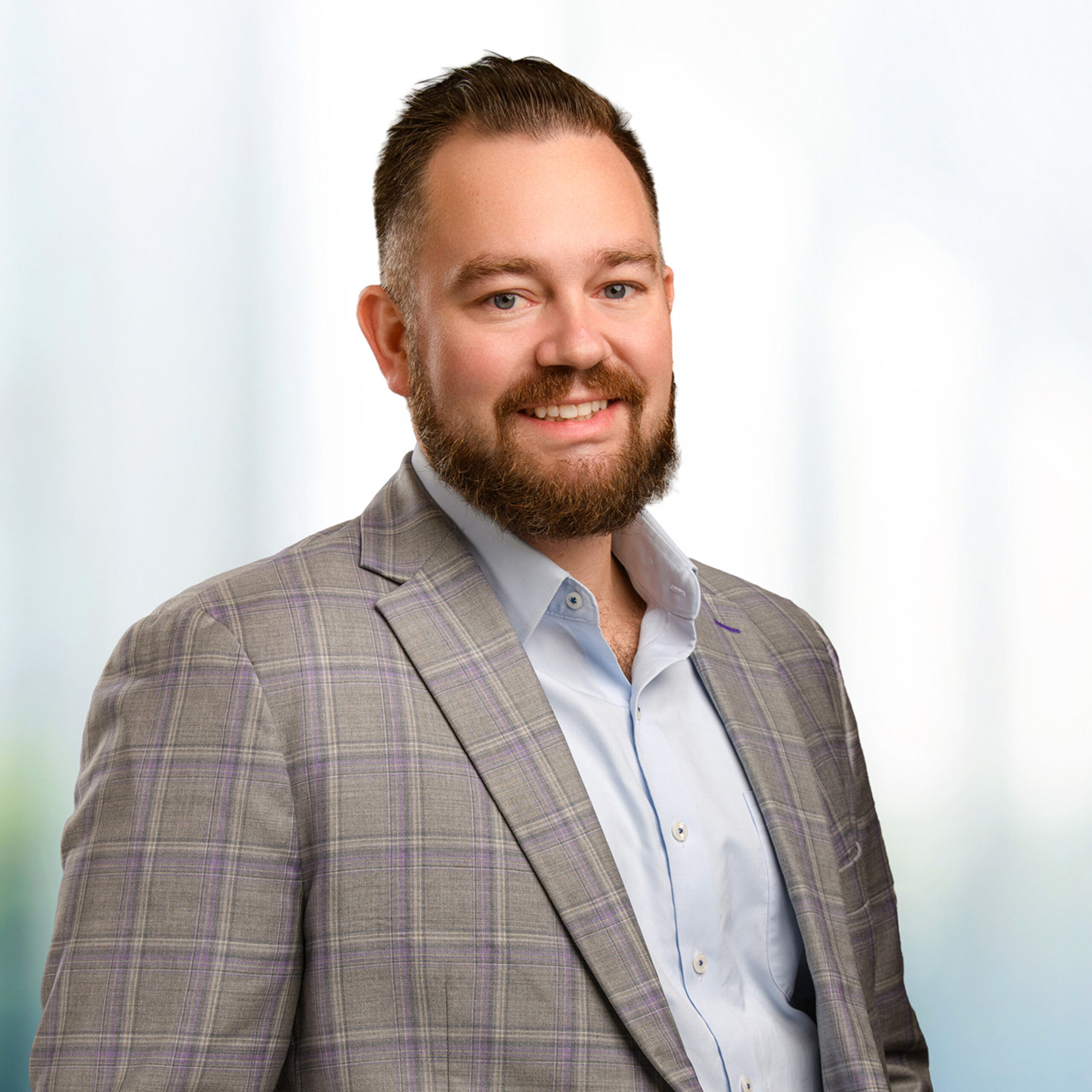 Smiling bearded man wearing a gray plaid suit jacket and light blue shirt against a soft blurred background.