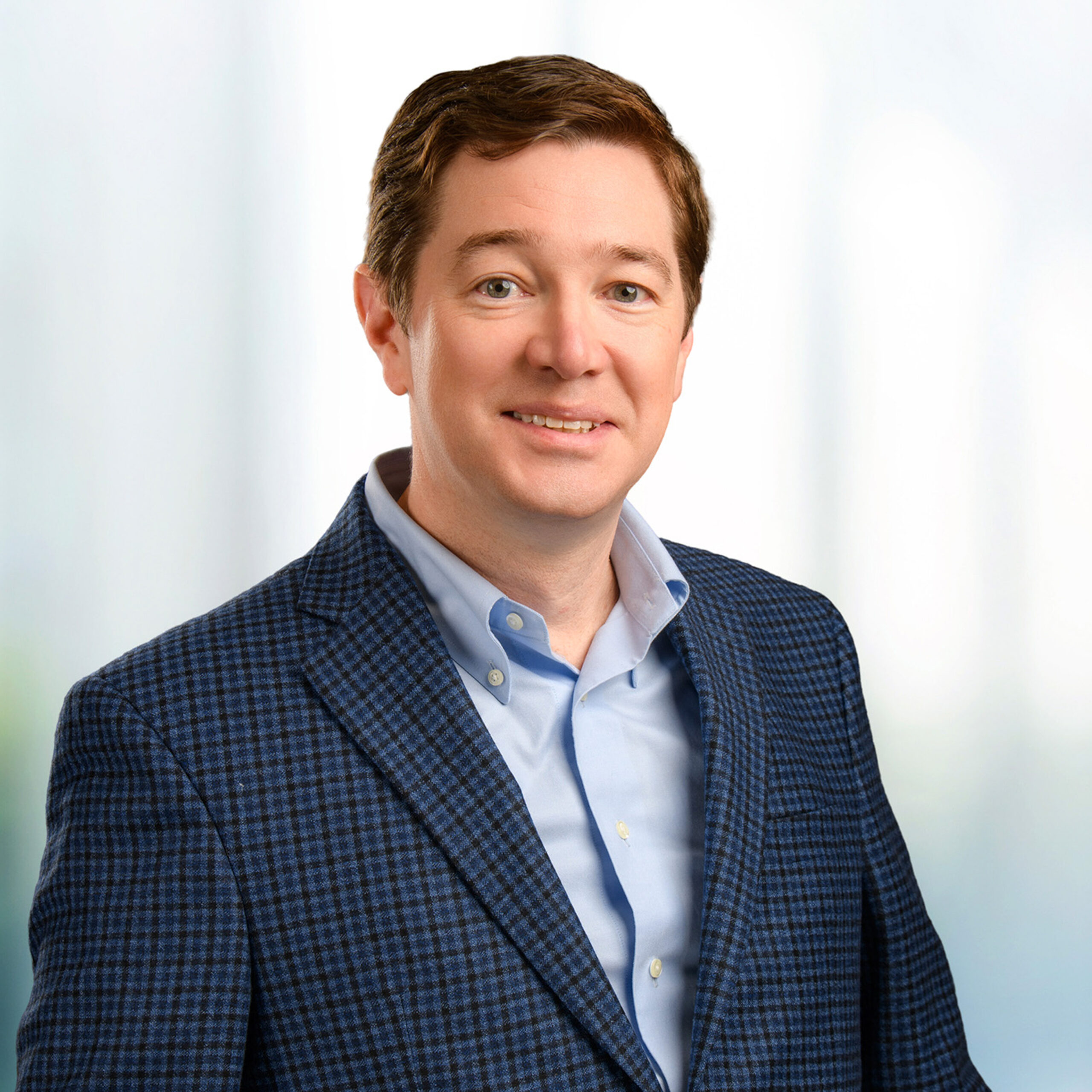 Smiling man with short brown hair wearing a blue checked blazer and light blue shirt against a blurred pale background.