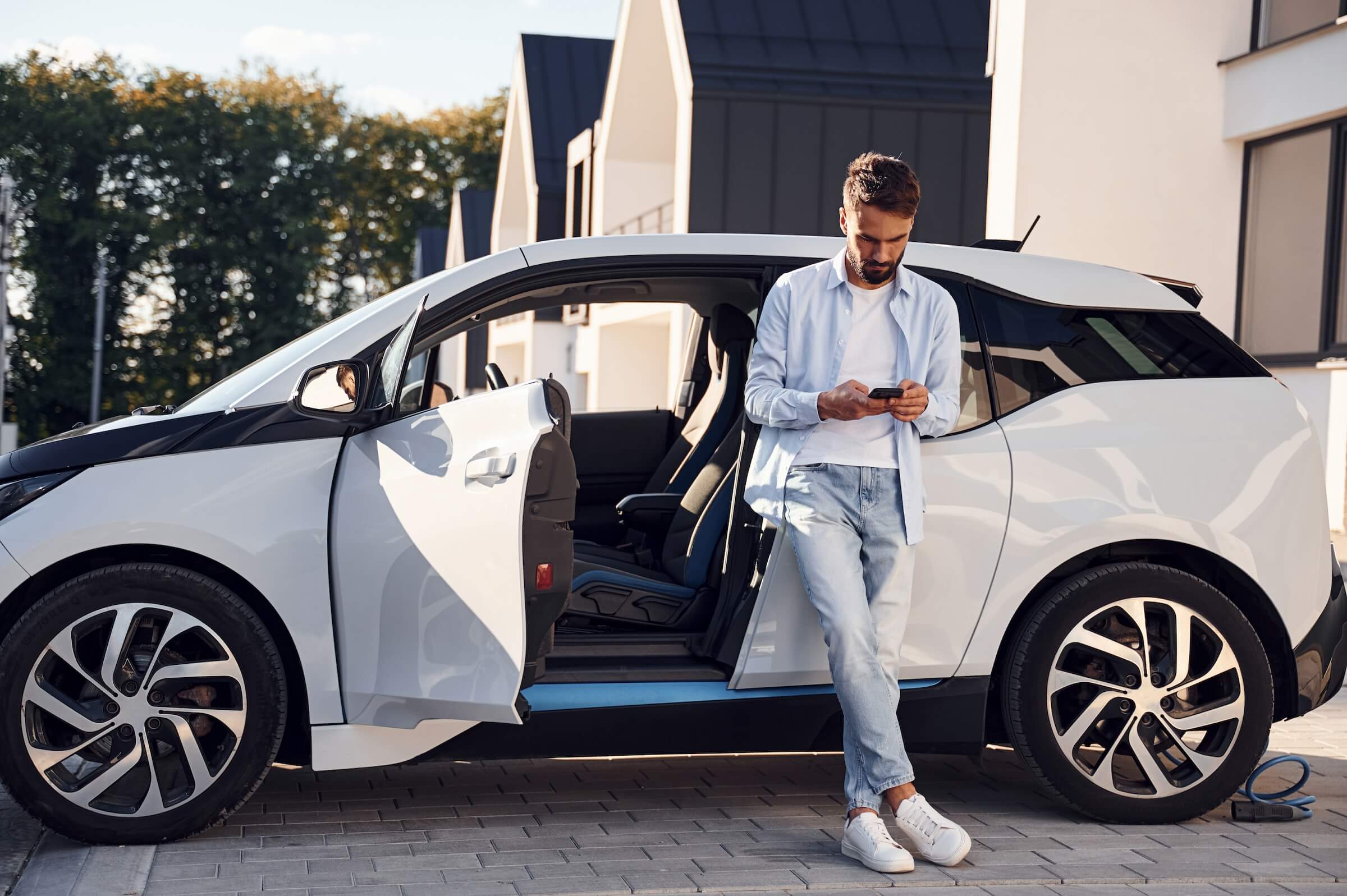Man in a light shirt and jeans leans against the open driver-side door of a white electric car parked in front of modern townhouses, looking at his phone while a charging cable lies coiled near the rear wheel.