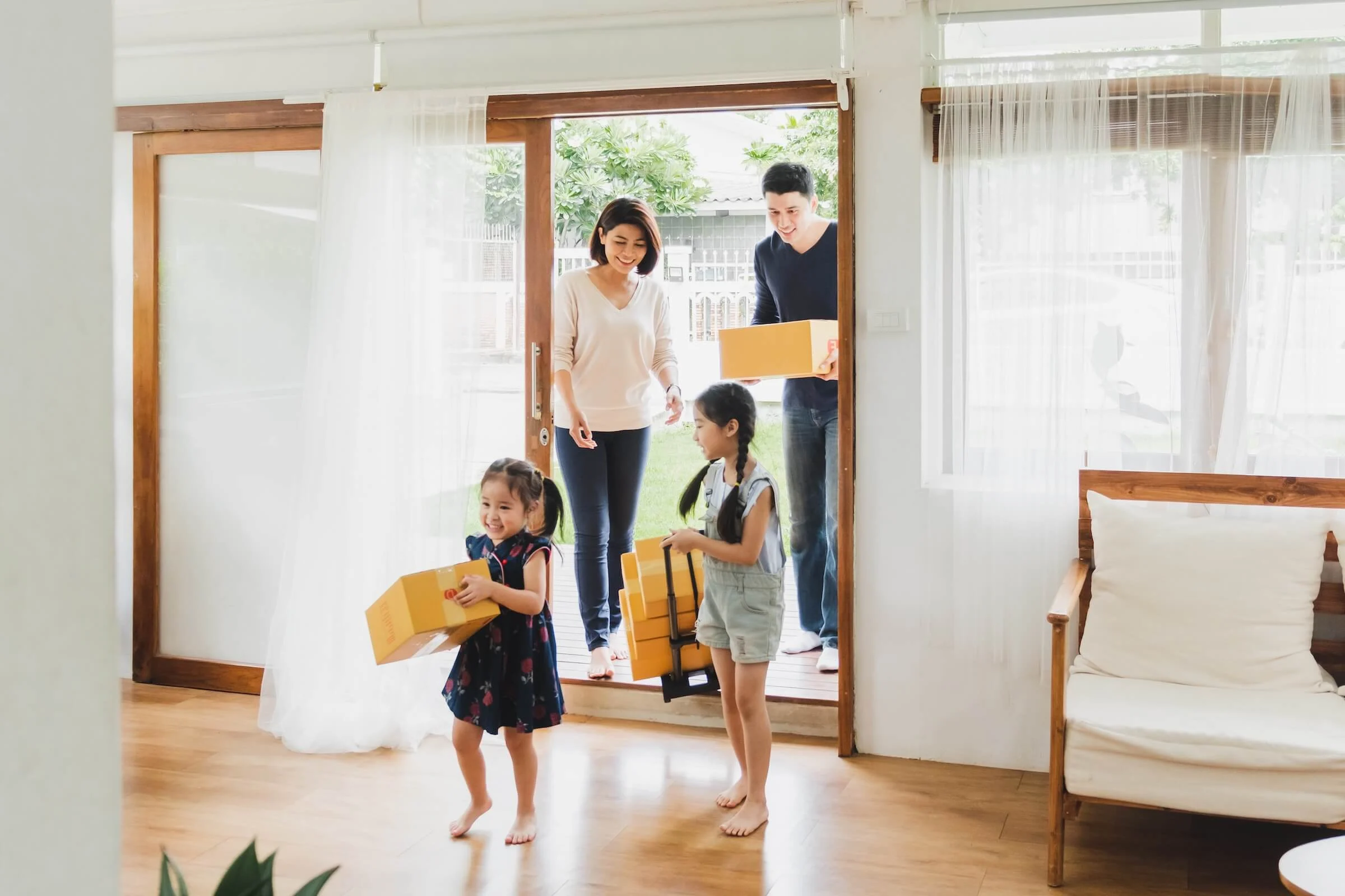 Two young girls carry yellow delivery boxes into a bright living room as their smiling parents stand in the open doorway holding more packages.