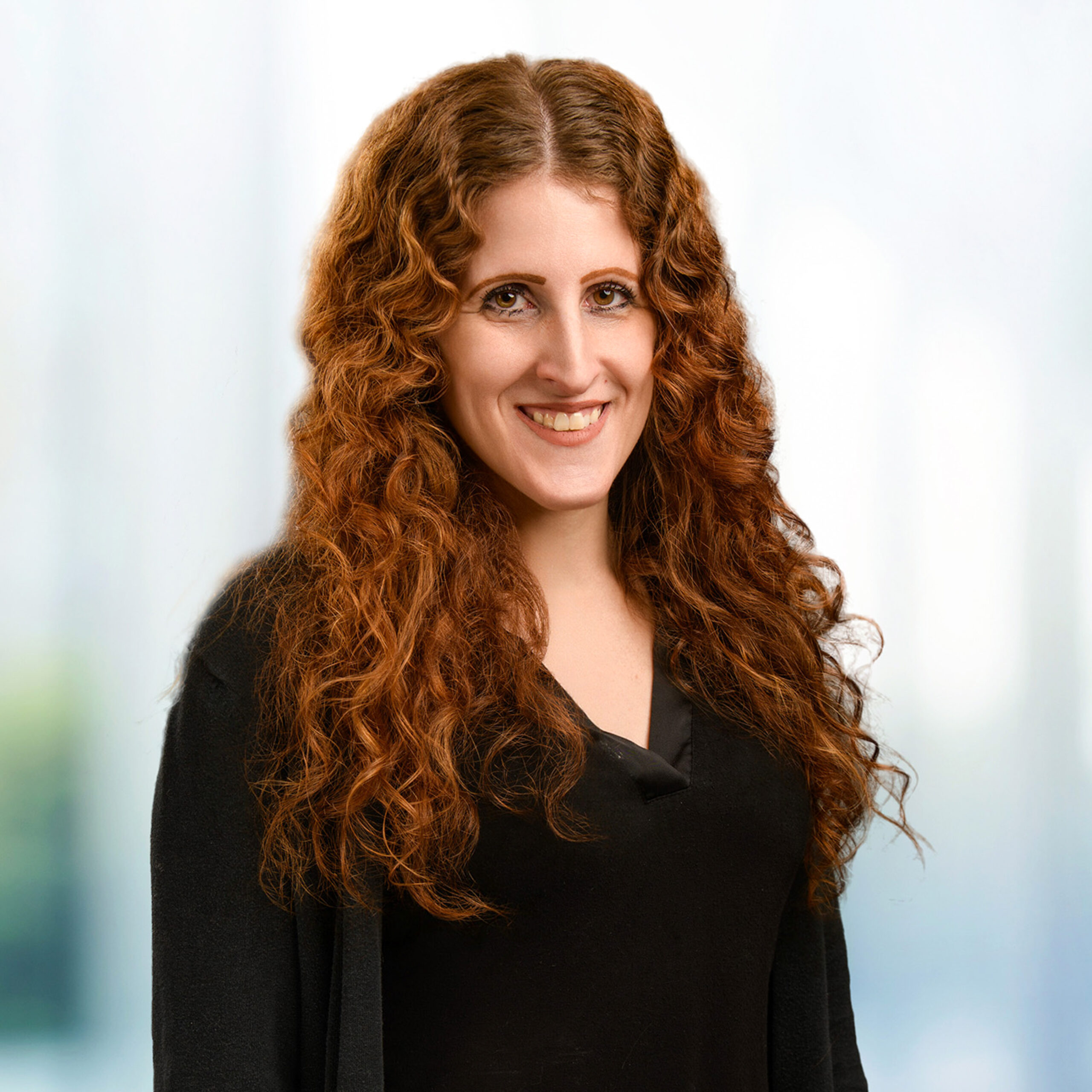 Woman with long curly auburn hair and warm smile wearing a black top, headshot against a soft blurred background.