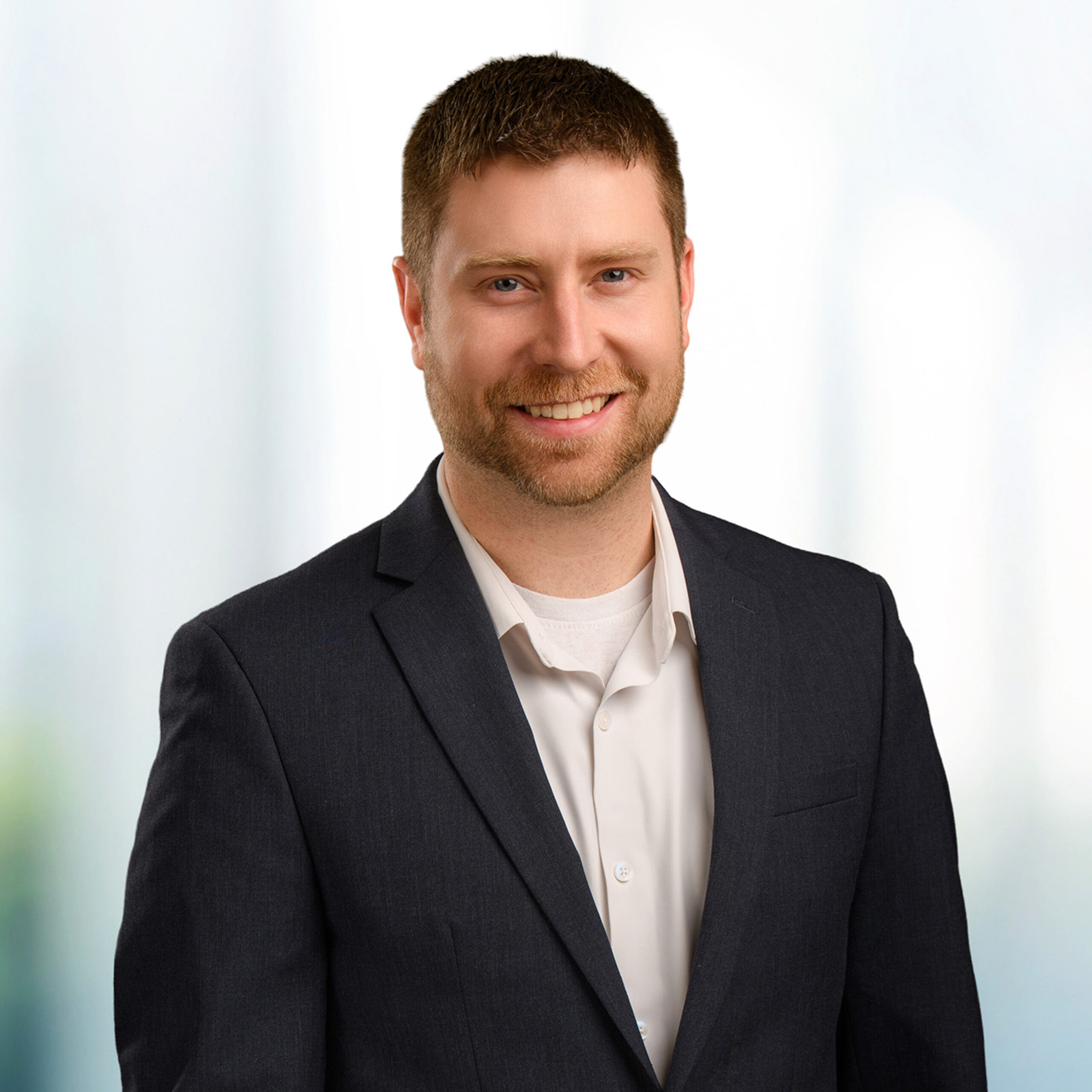 Smiling man with short brown hair and trimmed beard wearing a dark suit jacket and white shirt in a bright blurred background