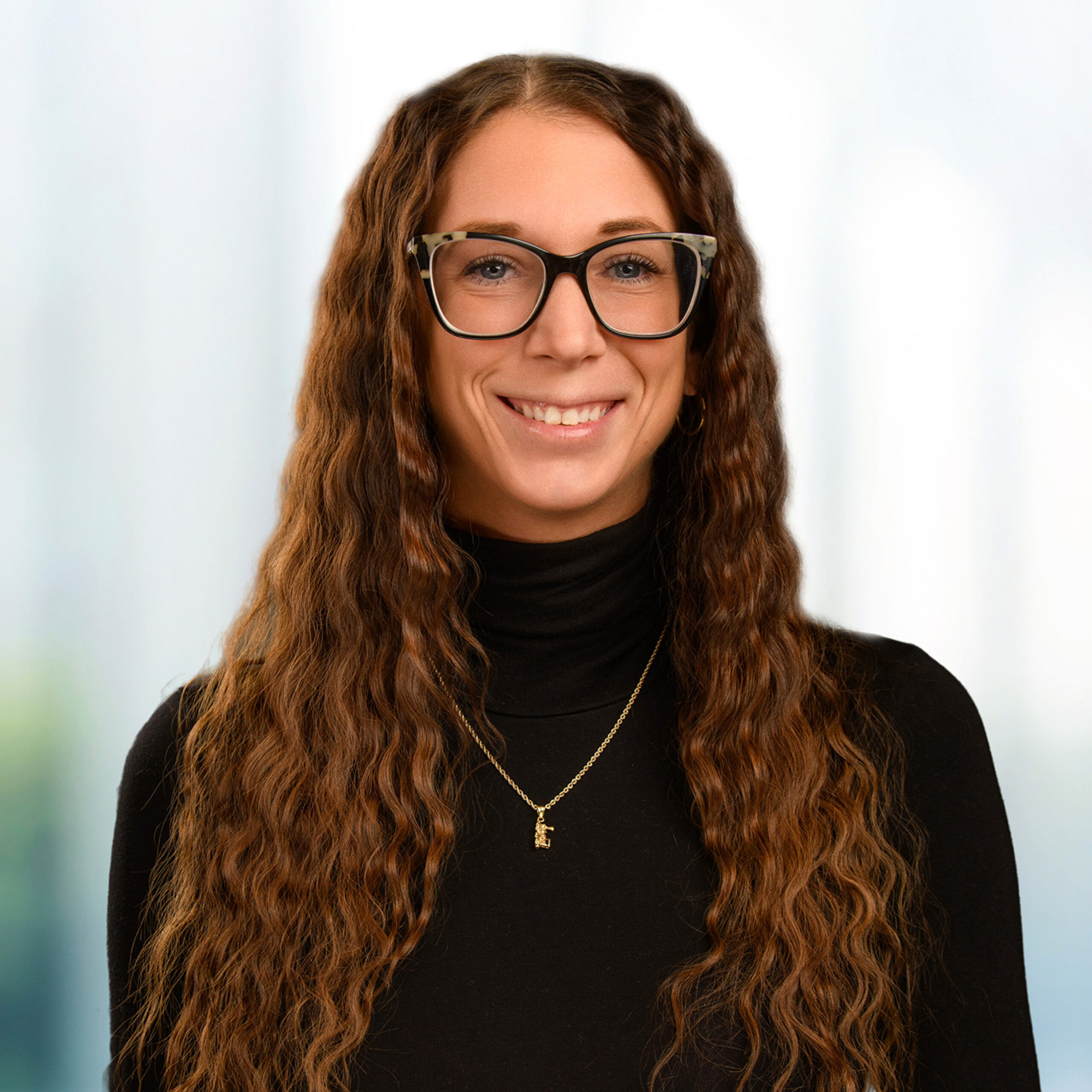 Smiling woman with long wavy brown hair wearing large tortoiseshell glasses, black turtleneck, and a gold pendant necklace.