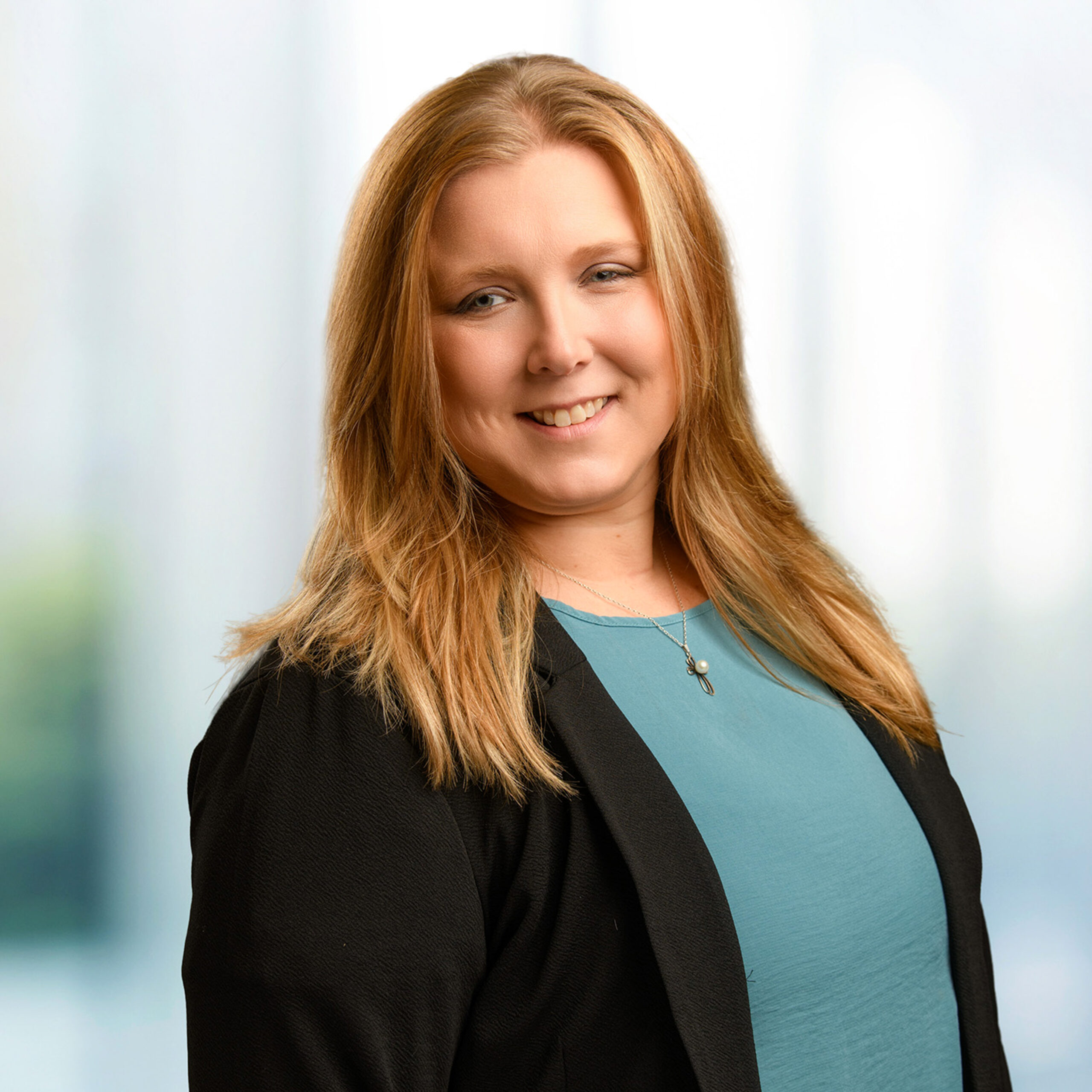 Smiling woman with long reddish-blonde hair in a teal top and black blazer, wearing a pendant necklace.