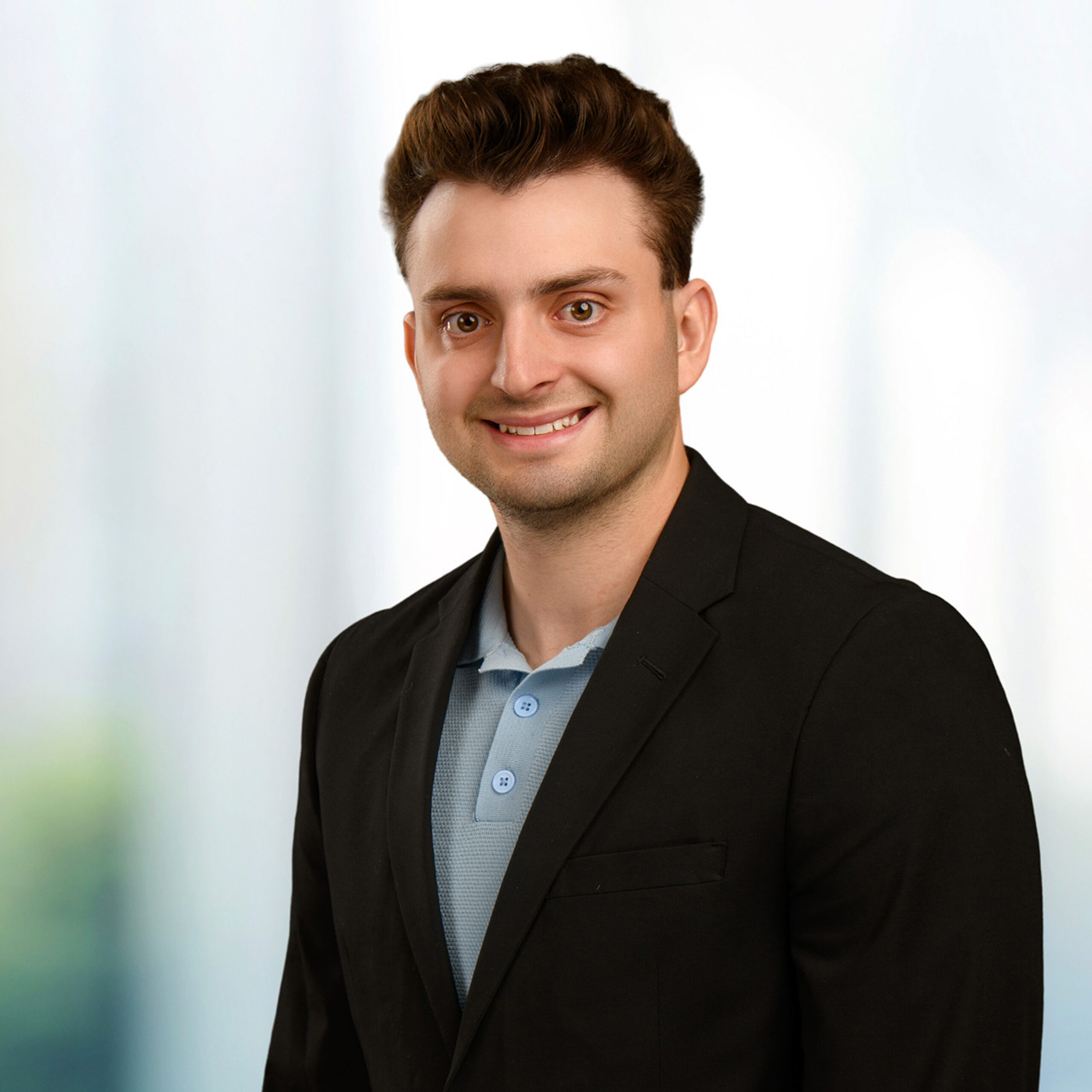 Young man with short brown hair smiling, wearing a black blazer over a light-blue polo against a blurred light background.
