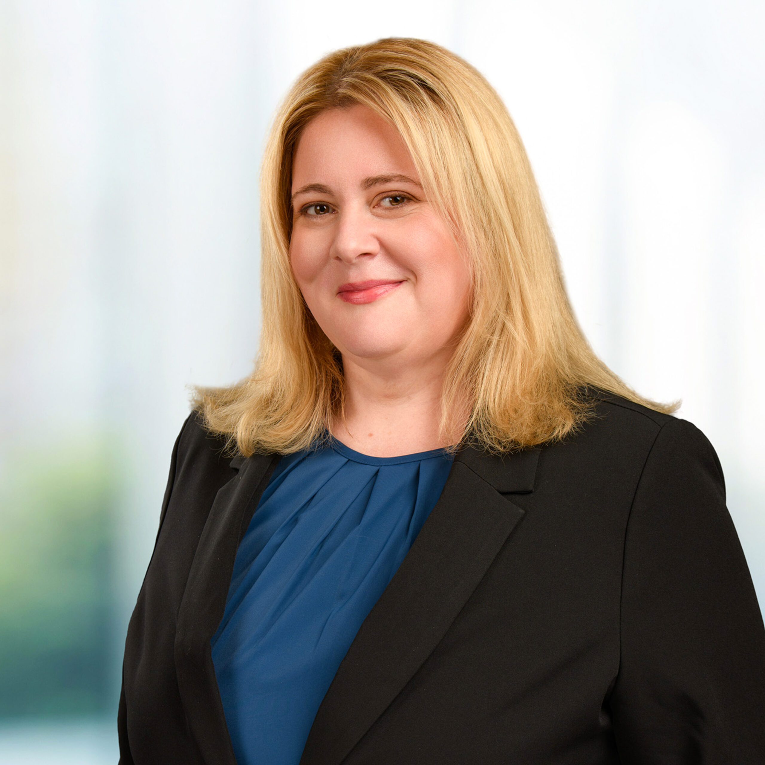 Professional headshot of a smiling blonde woman wearing a black blazer and blue blouse against a blurred background.