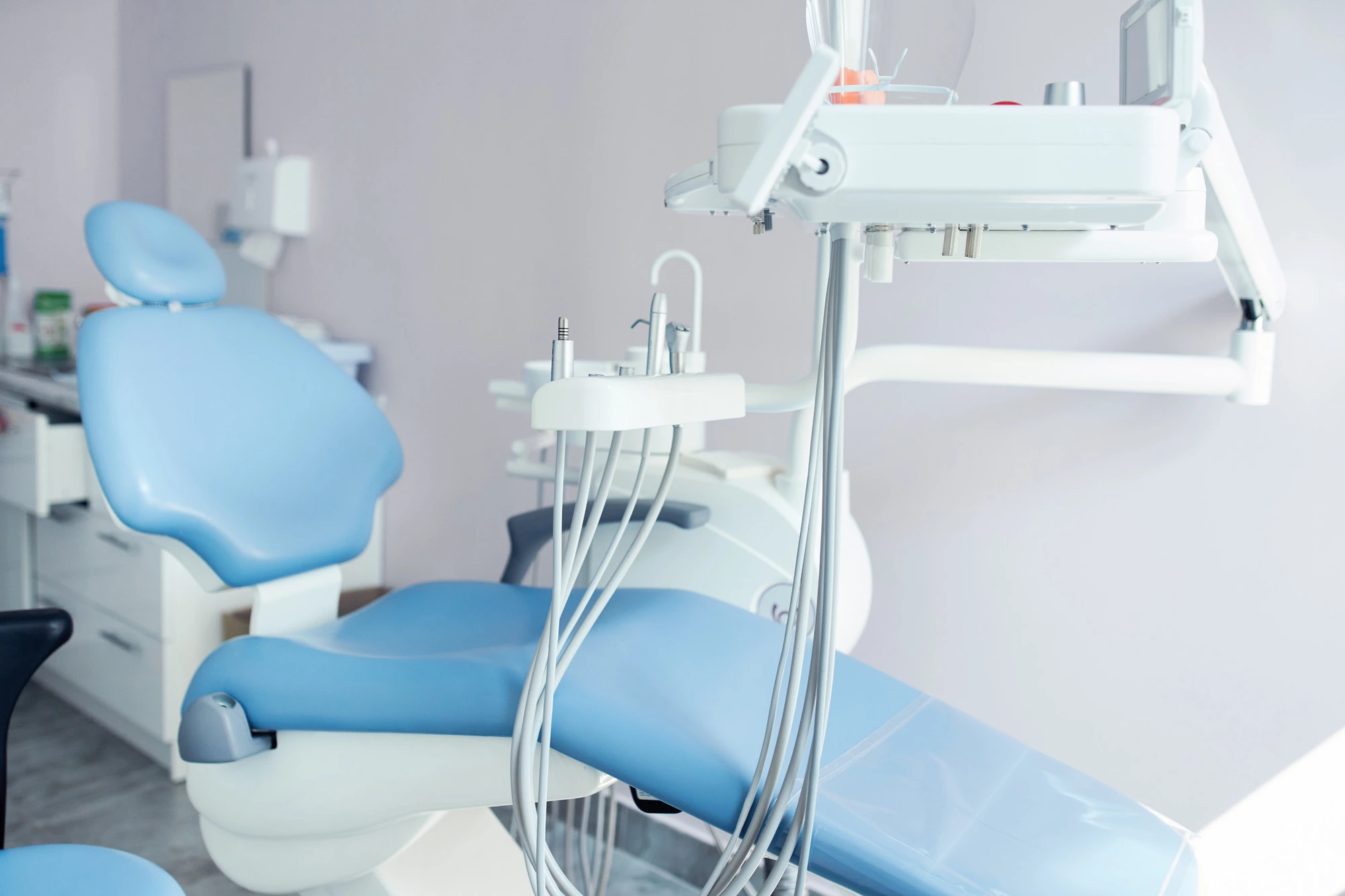 Modern blue dental chair in a bright treatment room with a white attached dental unit, instrument tray and hanging handpiece hoses.