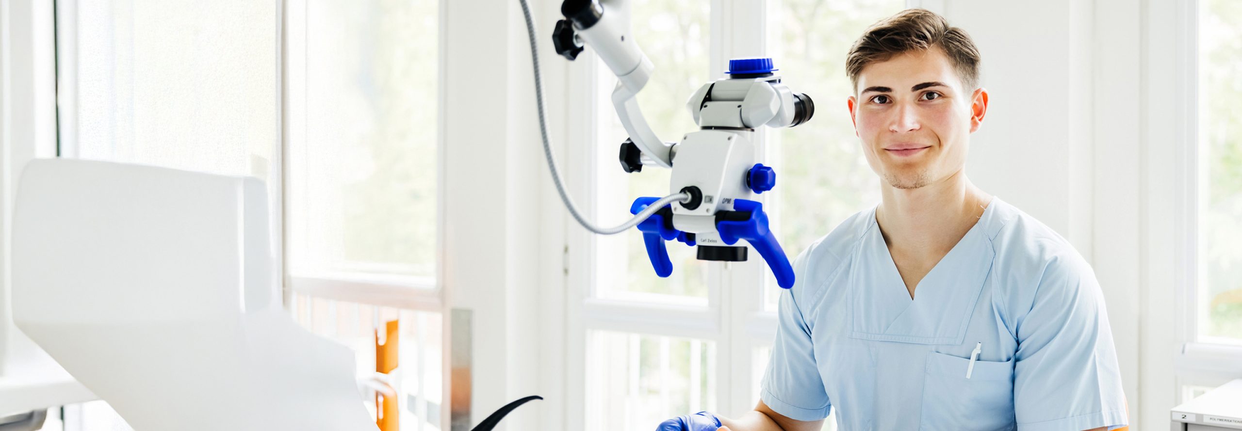 Young male dental professional in light-blue scrubs seated next to a white dental microscope with blue handles in a bright, modern treatment room.