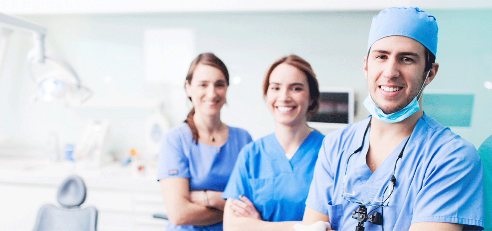 Three smiling dental professionals in blue scrubs in a bright clinic; a male dentist at right wears a surgical cap, face mask pulled down and dental loupes, with two colleagues standing behind near a dental chair and overhead lamp.