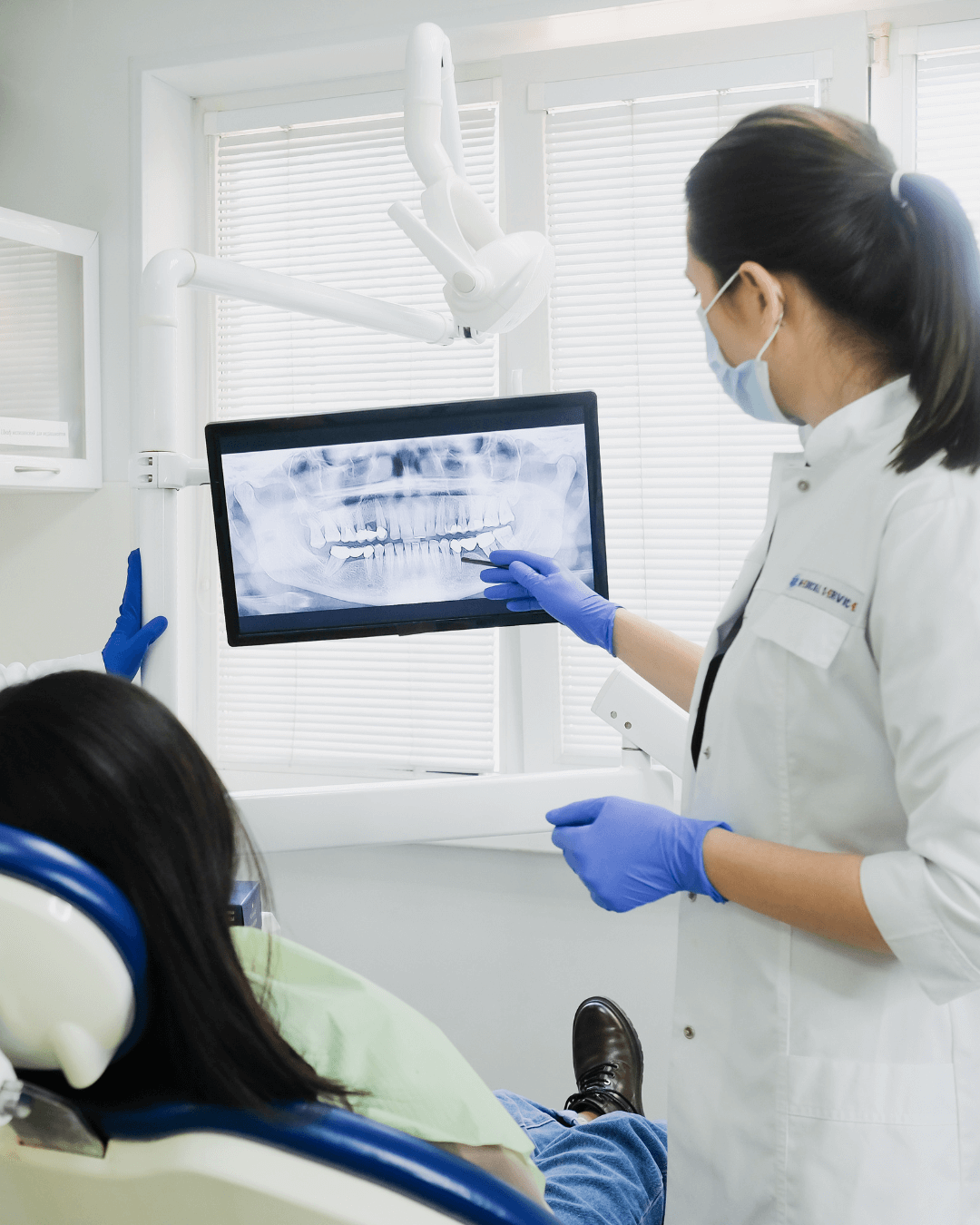 A dentist in a white coat wearing a mask and blue gloves points to a panoramic dental X-ray displayed on a monitor while a patient reclines in the dental chair.