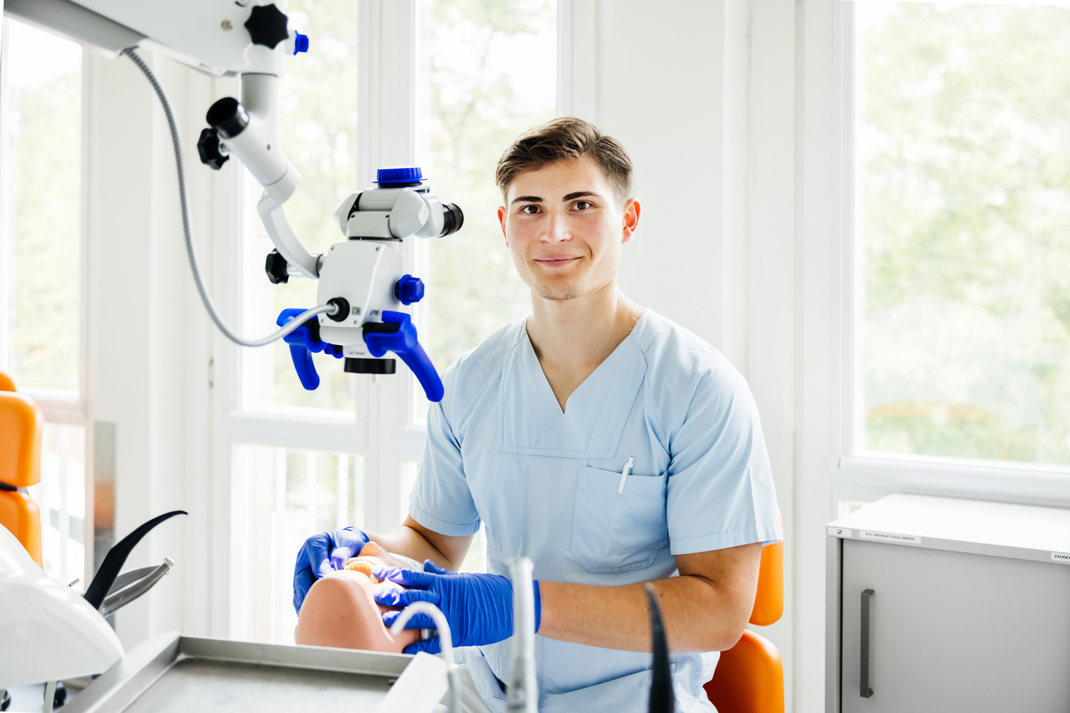 Young dental professional in light-blue scrubs and blue gloves smiling at the camera while working on a dental training mannequin under a white-and-blue dental microscope in a bright treatment room with large windows.